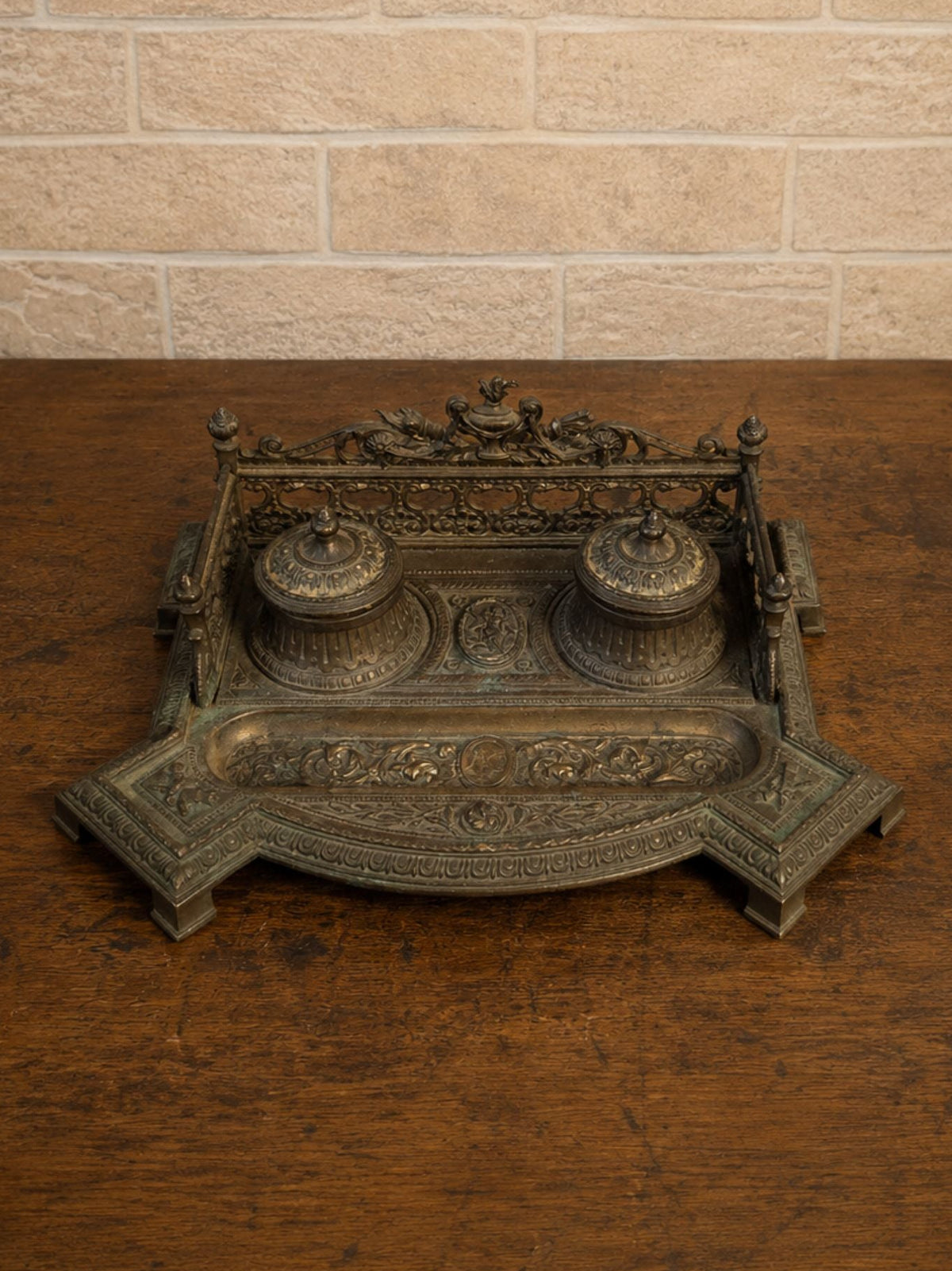 An ornate antique-style bronze desk organizer with two small, lidded inkwells and a decorative top frame, displayed on a dark wooden table against a light brick wall.