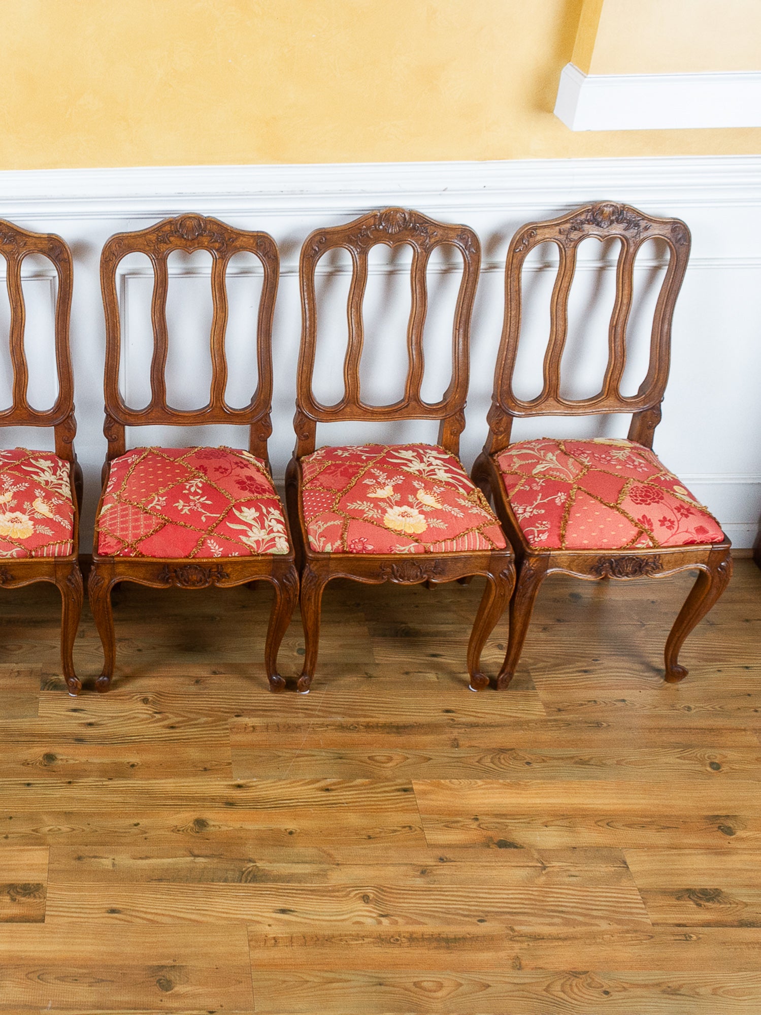 Four vintage wooden dining chairs with carved backs and red floral patterned upholstery, arranged on a wooden floor against a yellow wall.
