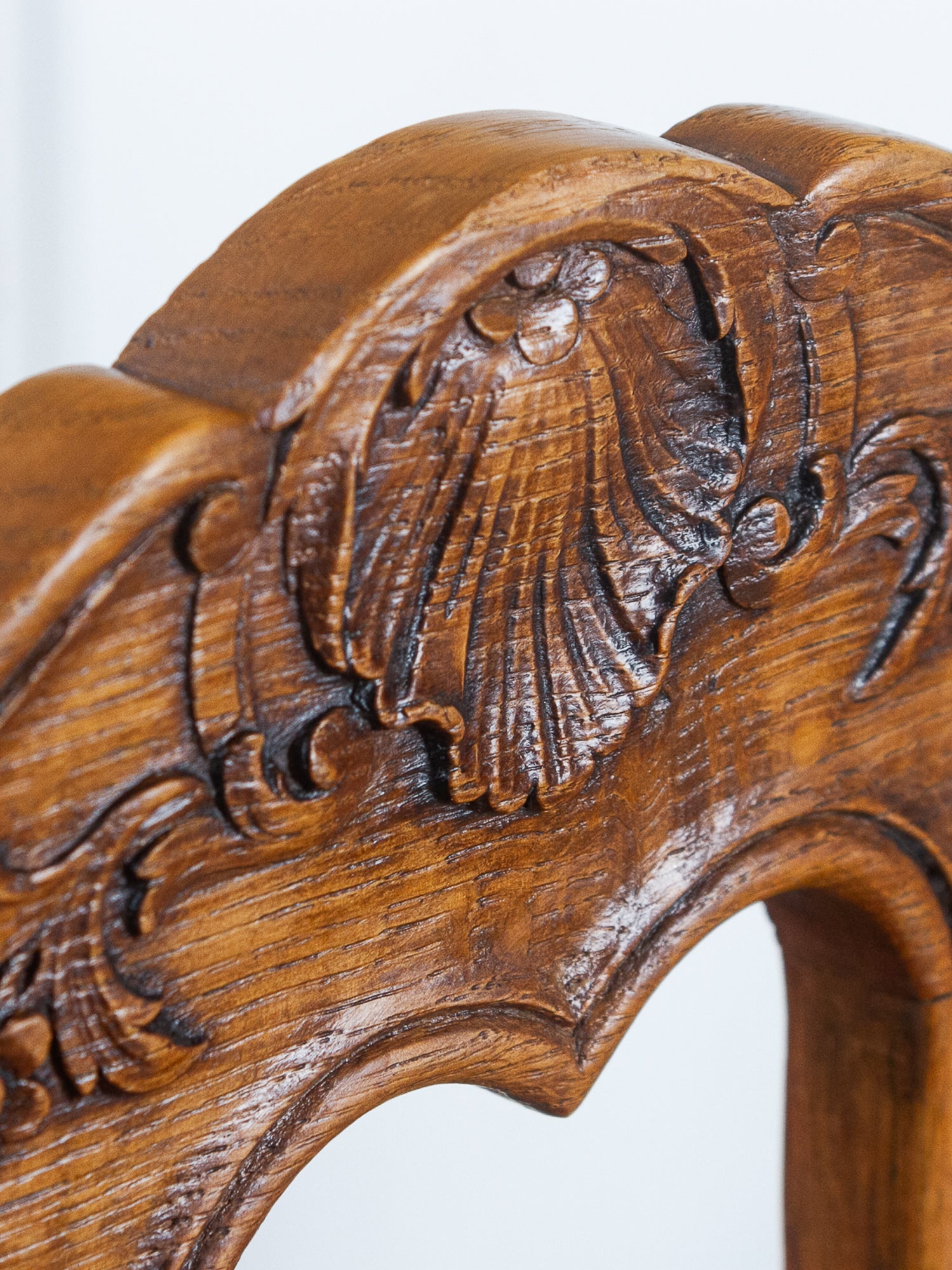 Close-up of an ornate carved wooden chair back or frame, showing rich medium-brown wood grain and detailed scrollwork against a light neutral background.