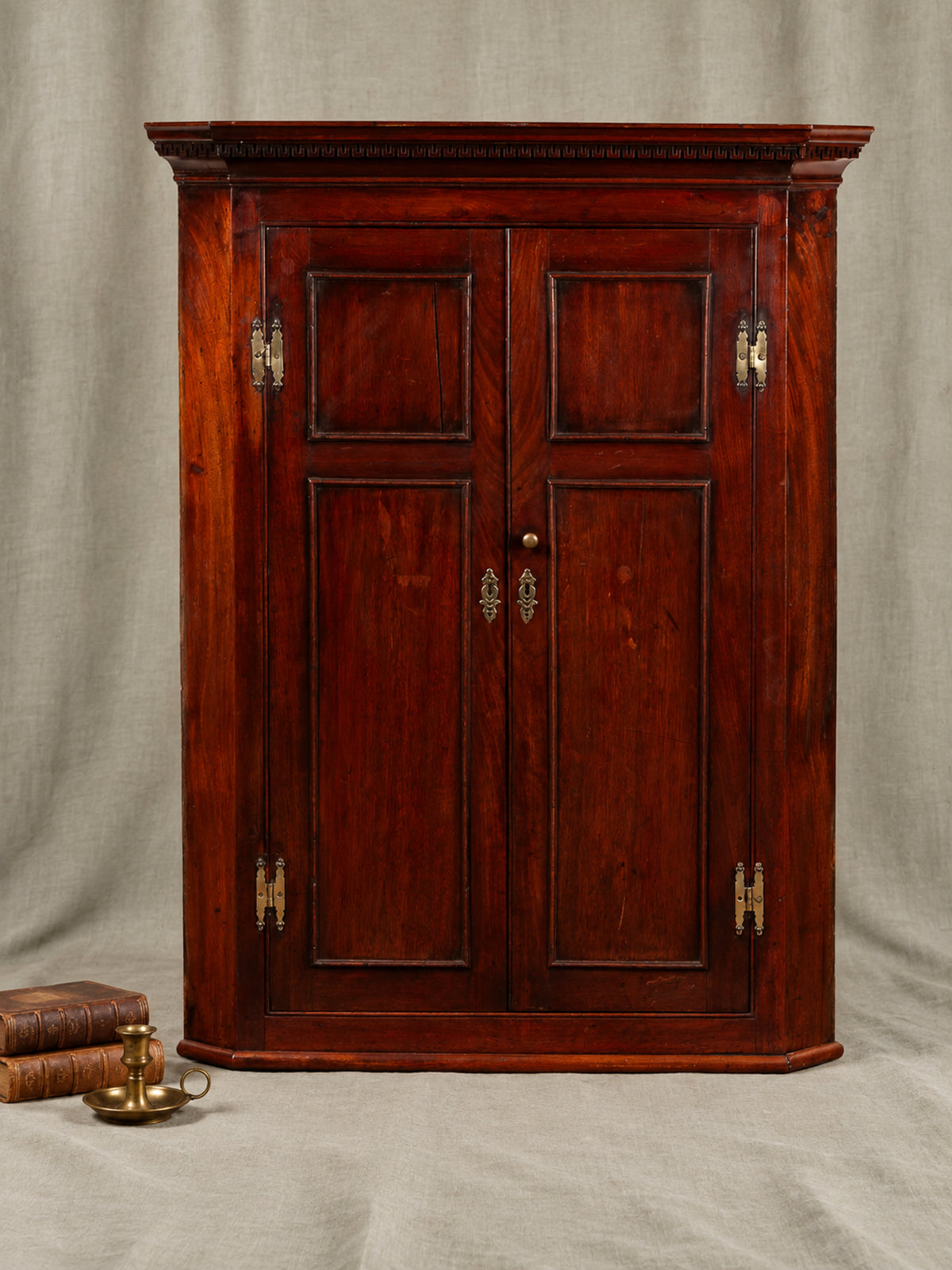 A vintage dark reddish-brown Mahogany  wooden cabinet with two paneled doors and brass hinges, set against a neutral draped fabric backdrop with decorative books in the lower left corner.