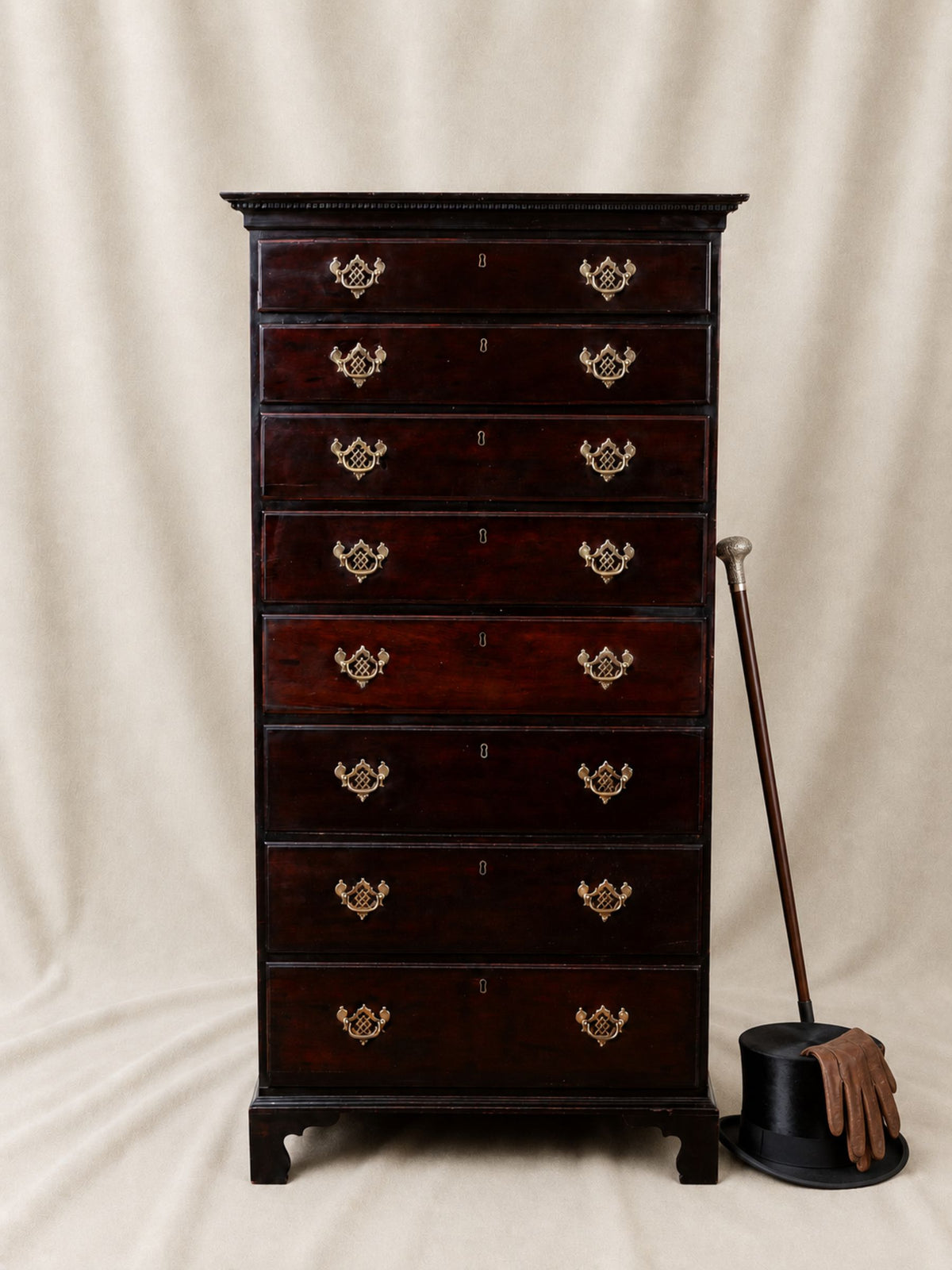A tall antique-style wooden chest of drawers with ten drawers, featuring ornate brass handles and a dark reddish-brown finish, photographed against a draped beige fabric backdrop with a cane and top hat prop beside it.
