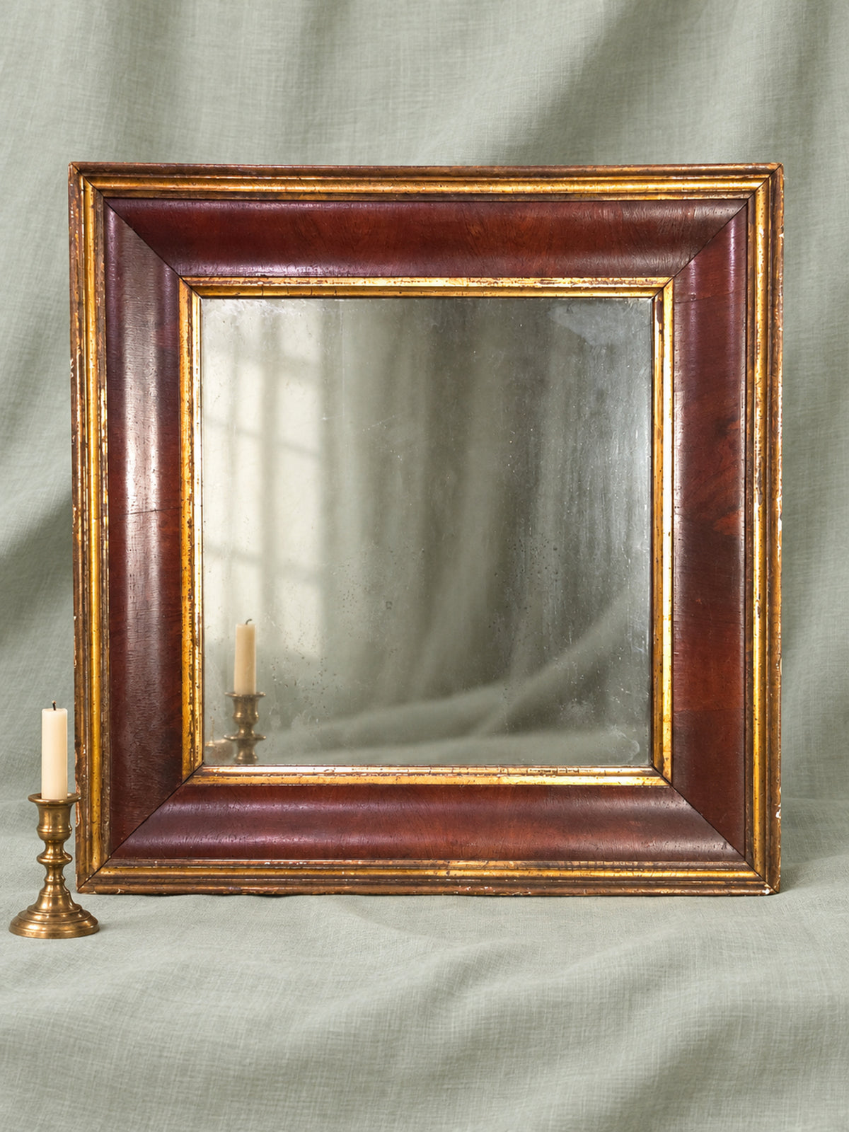 A square antique-style wall mirror with a dark reddish-brown wooden frame and distressed gold trim, set against a draped sage green fabric backdrop with a brass candlestick prop on the left.