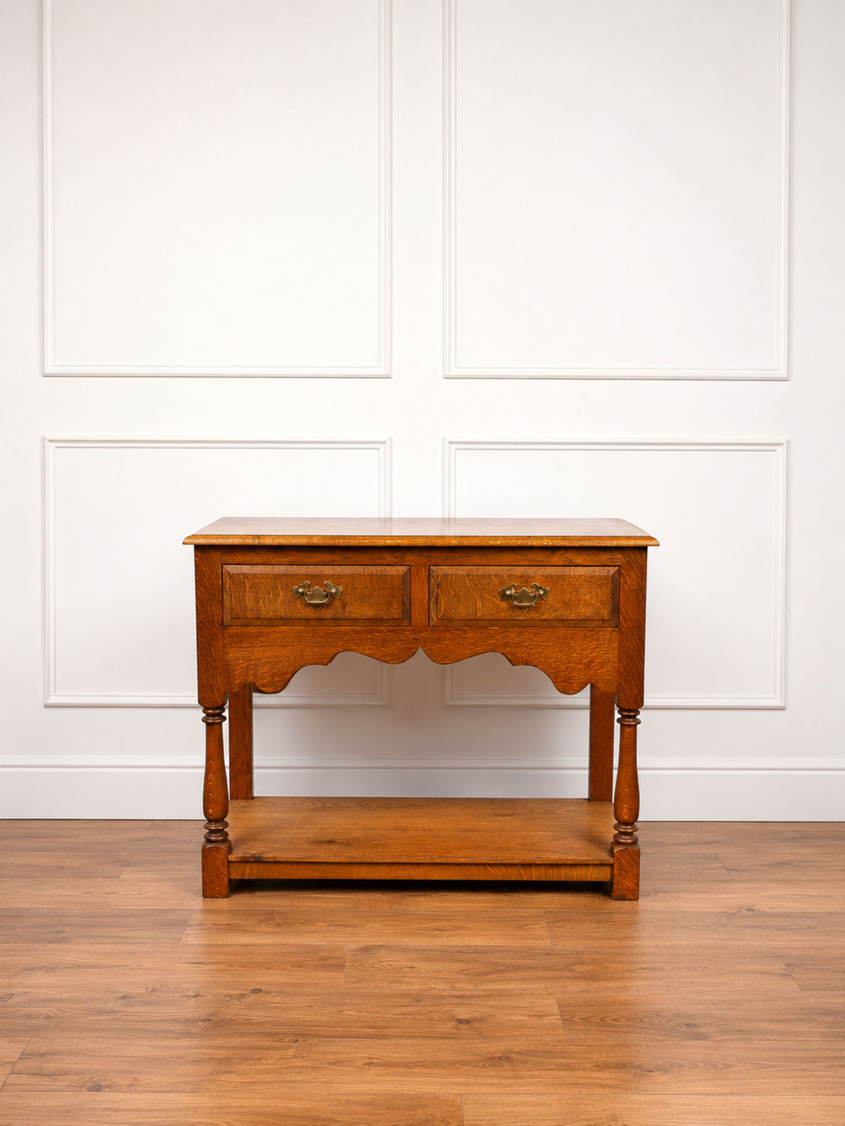 An antique oak wooden console table with two drawers, turned legs, and a lower shelf, set against a white paneled wall and hardwood floor.