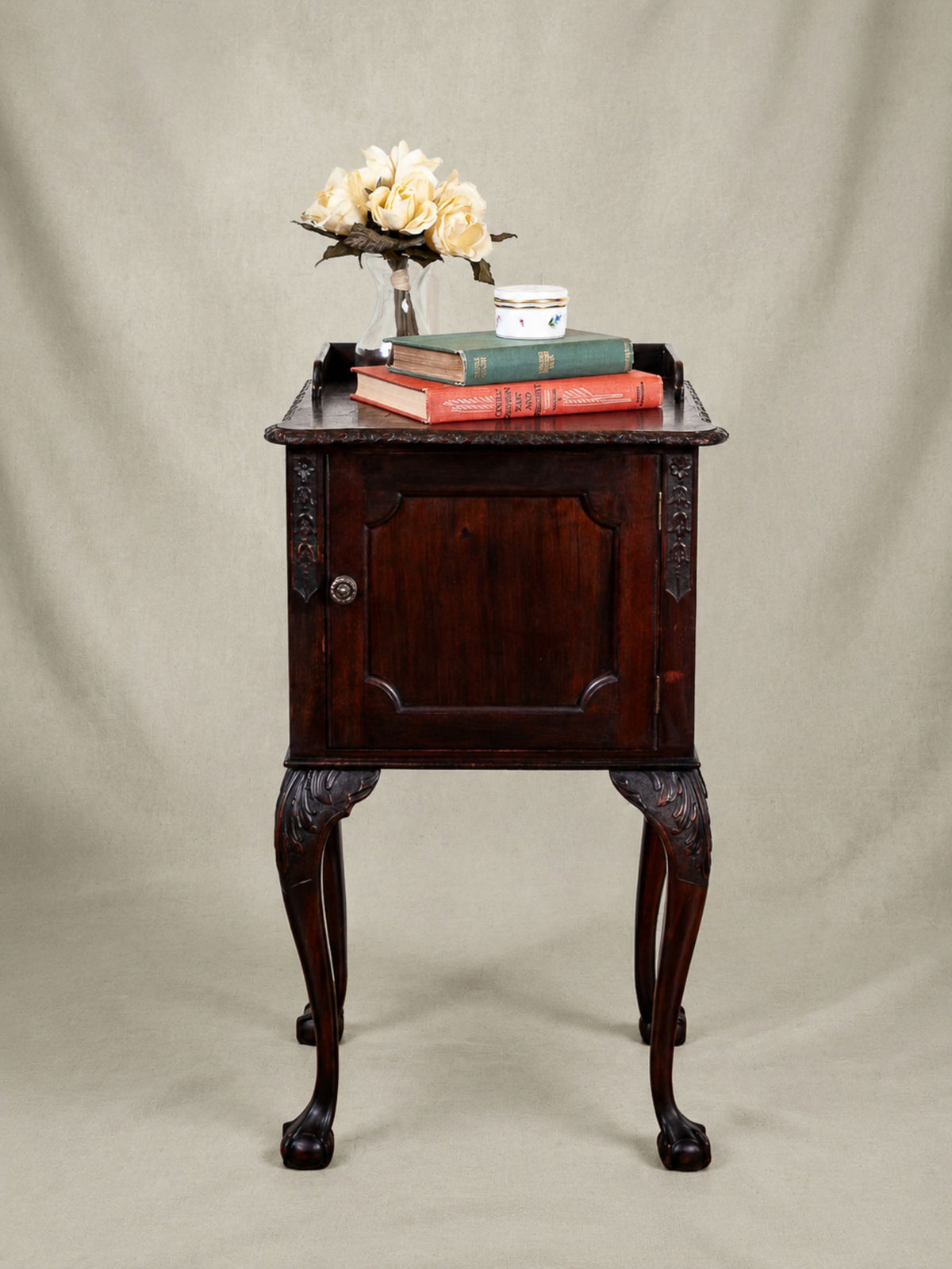 A dark English Mahogany carved wooden nightstand with cabriole legs, styled with books, a small vase of flowers, and a teacup against a neutral beige fabric backdrop.