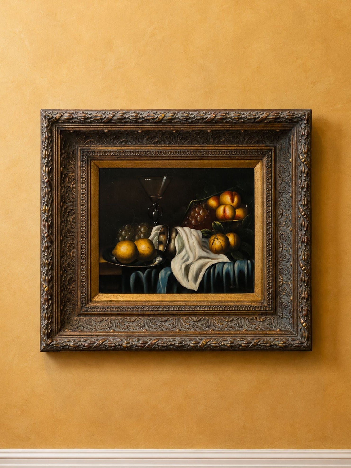 A framed still life painting of fruit and a draped cloth, displayed on a textured mustard-yellow wall above white baseboard and wood flooring.