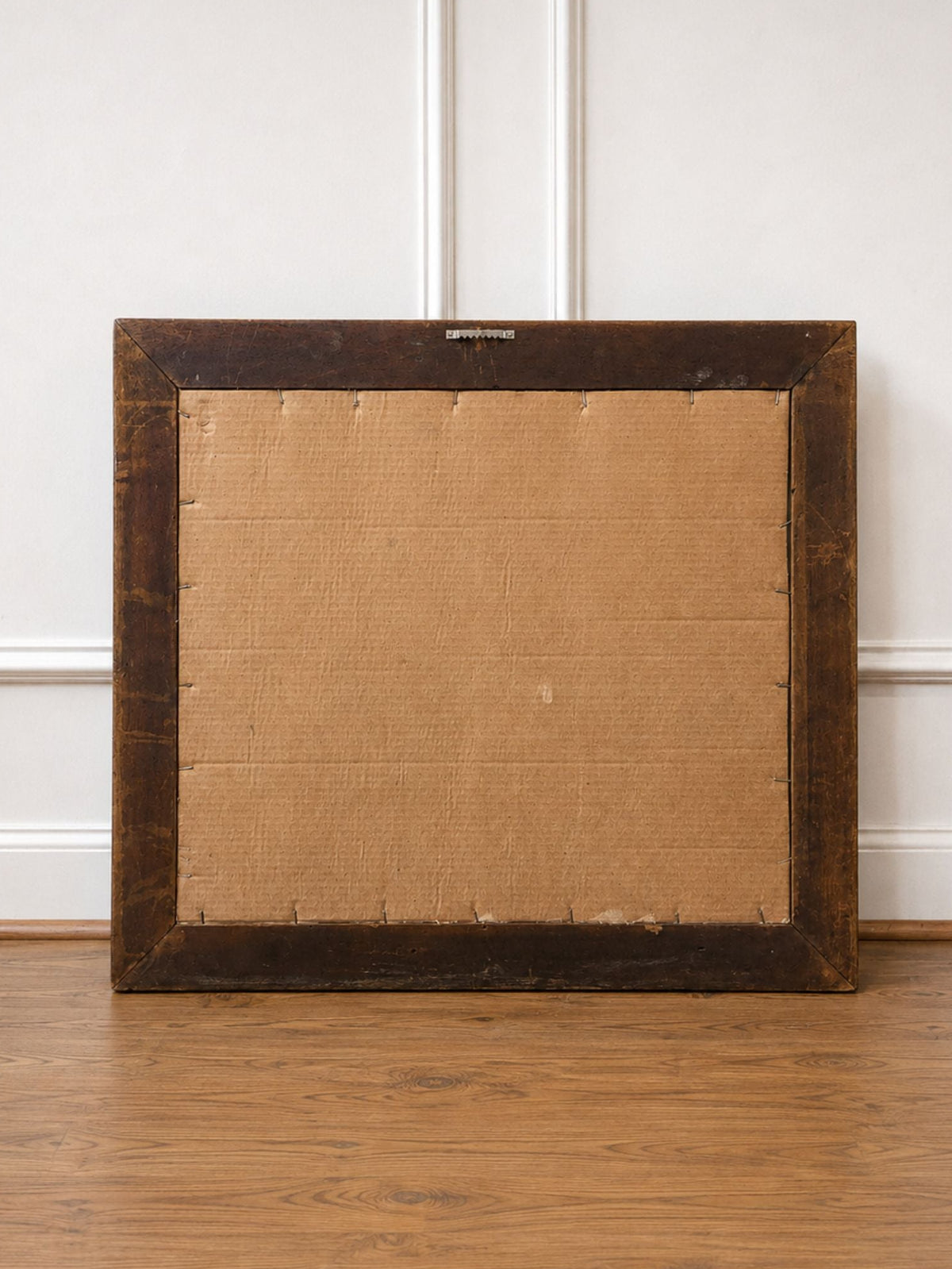 The back of a square framed artwork or panel, showing a dark brown wooden frame and tan canvas backing, standing on a wooden floor against a white paneled wall.