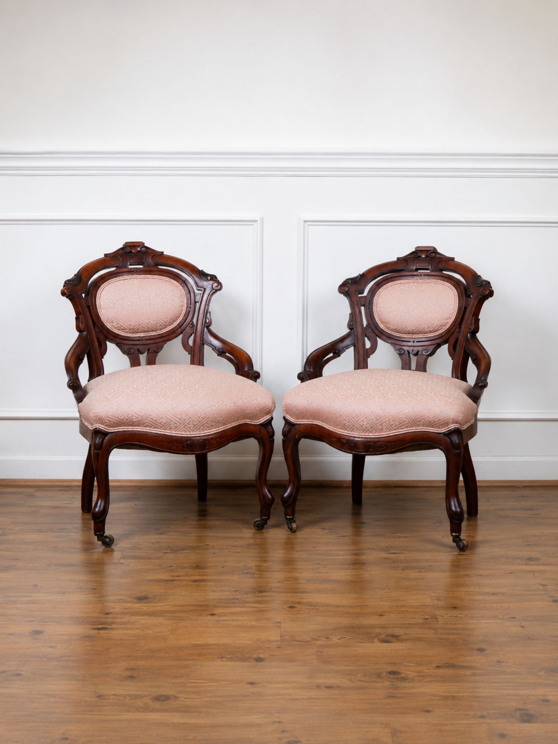 A pair of antique-style wooden armchairs with carved dark mahogany frames and dusty pink upholstered seats, set against a white paneled wall and hardwood floor.