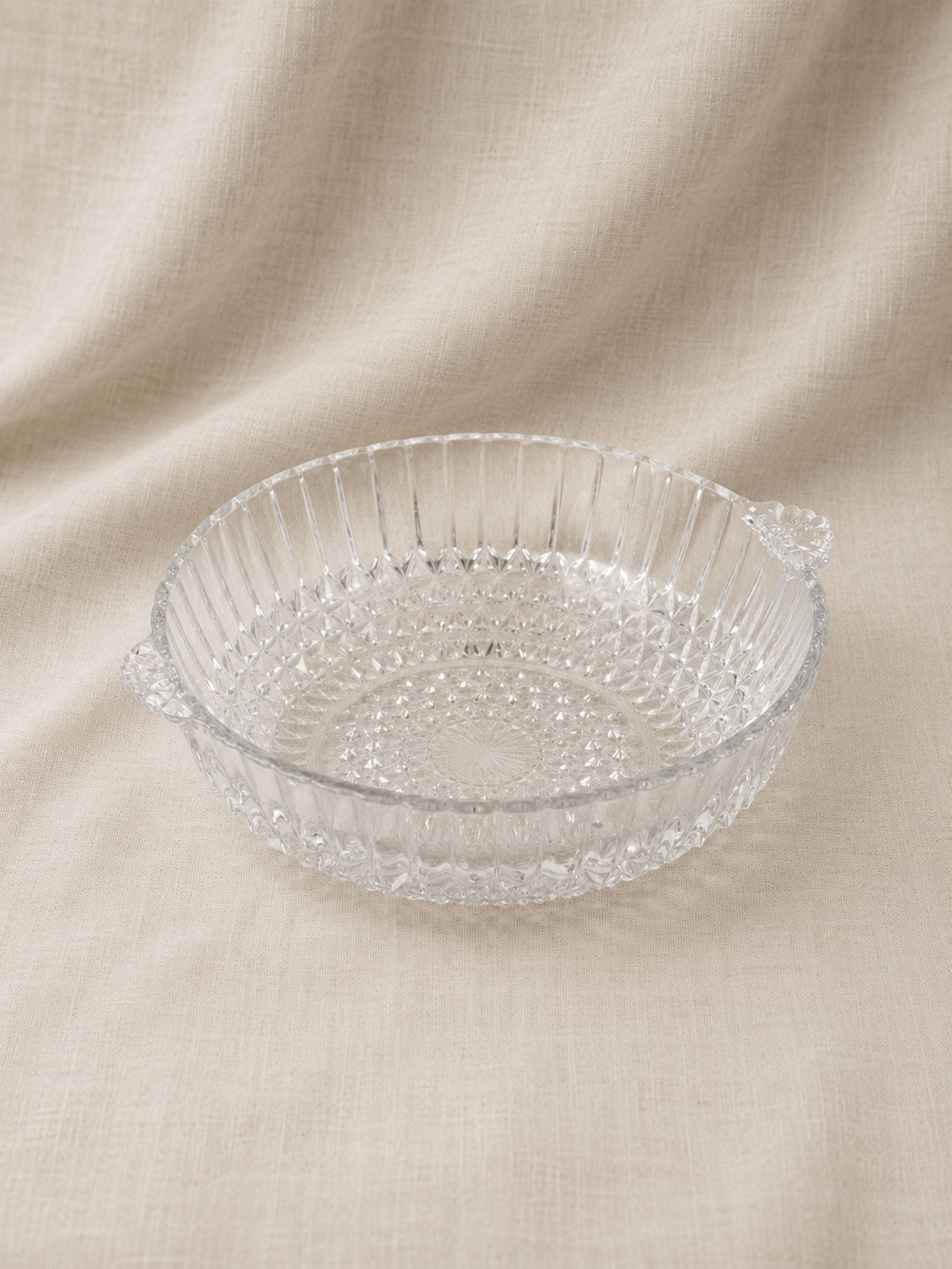 A clear glass baking dish with a textured, faceted pattern and small side handles, shown on a draped beige linen fabric background.