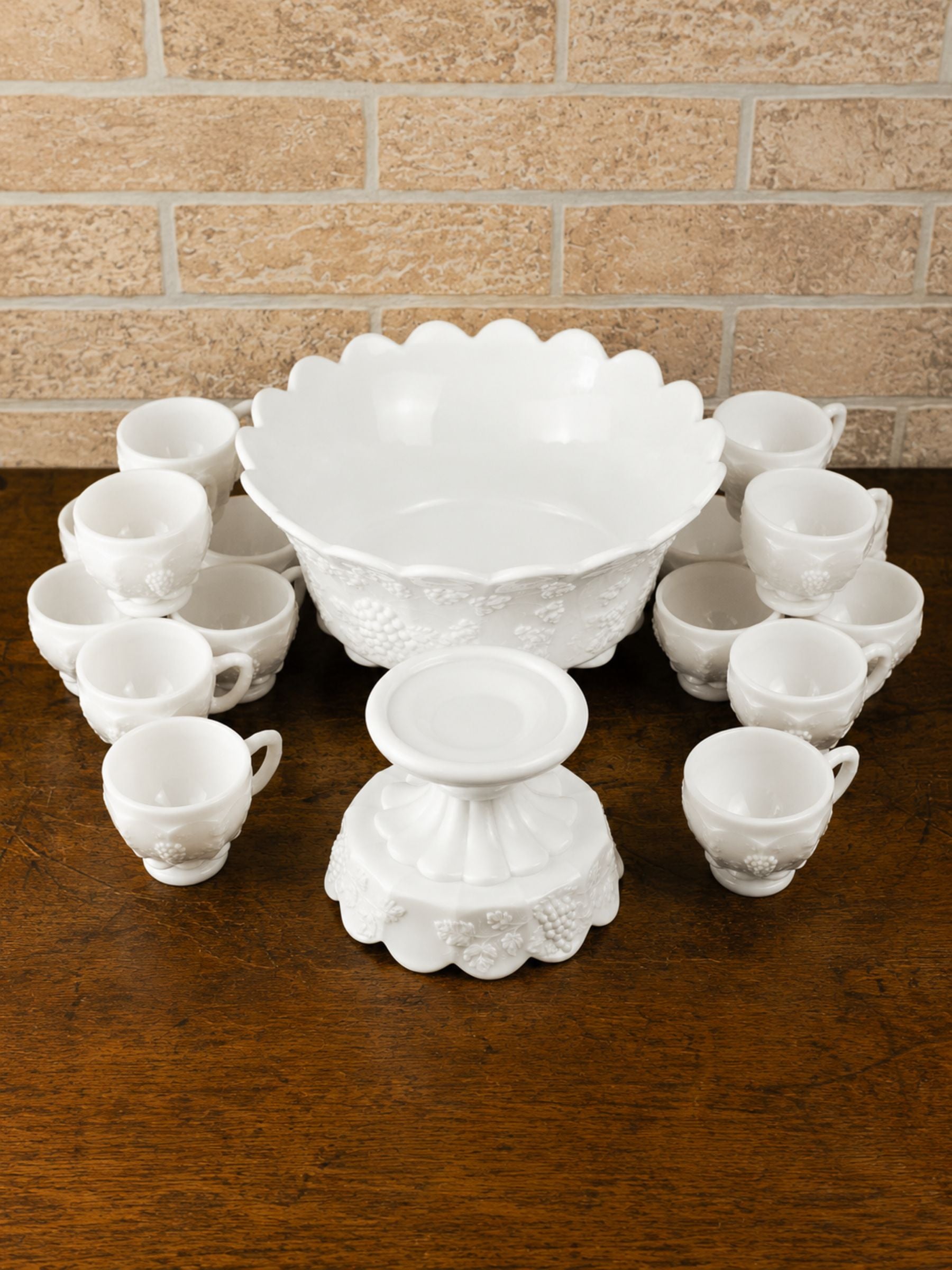 A white embossed glass party serving set arranged on a dark wooden table against a tan brick wall background. The set includes a large scalloped bowl, a pedestal base, a smaller bowl, and fourteen small handled cups.