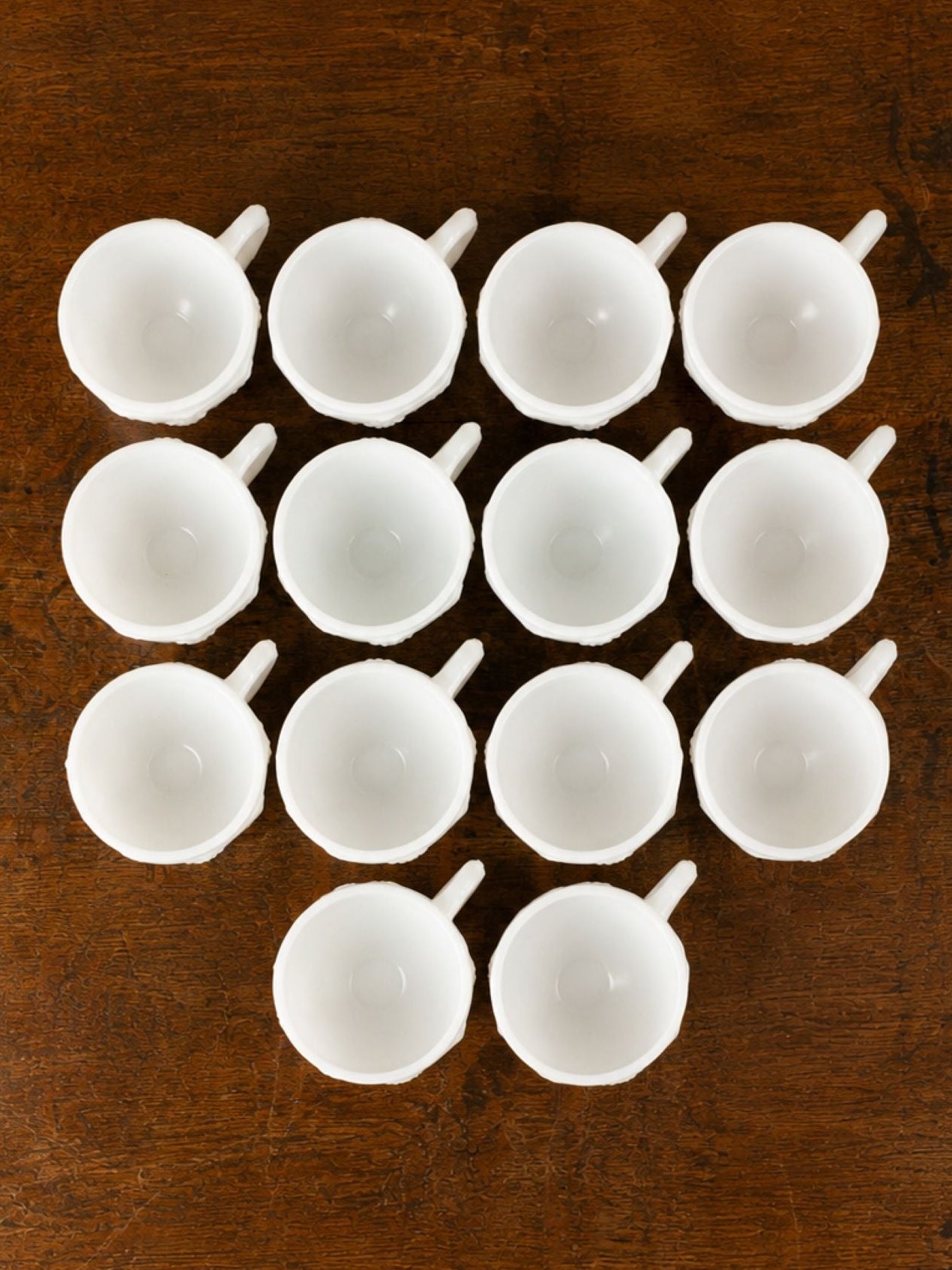 A set of 14 small milk glass cups arranged in a grid on a dark brown wooden table, viewed from directly above.