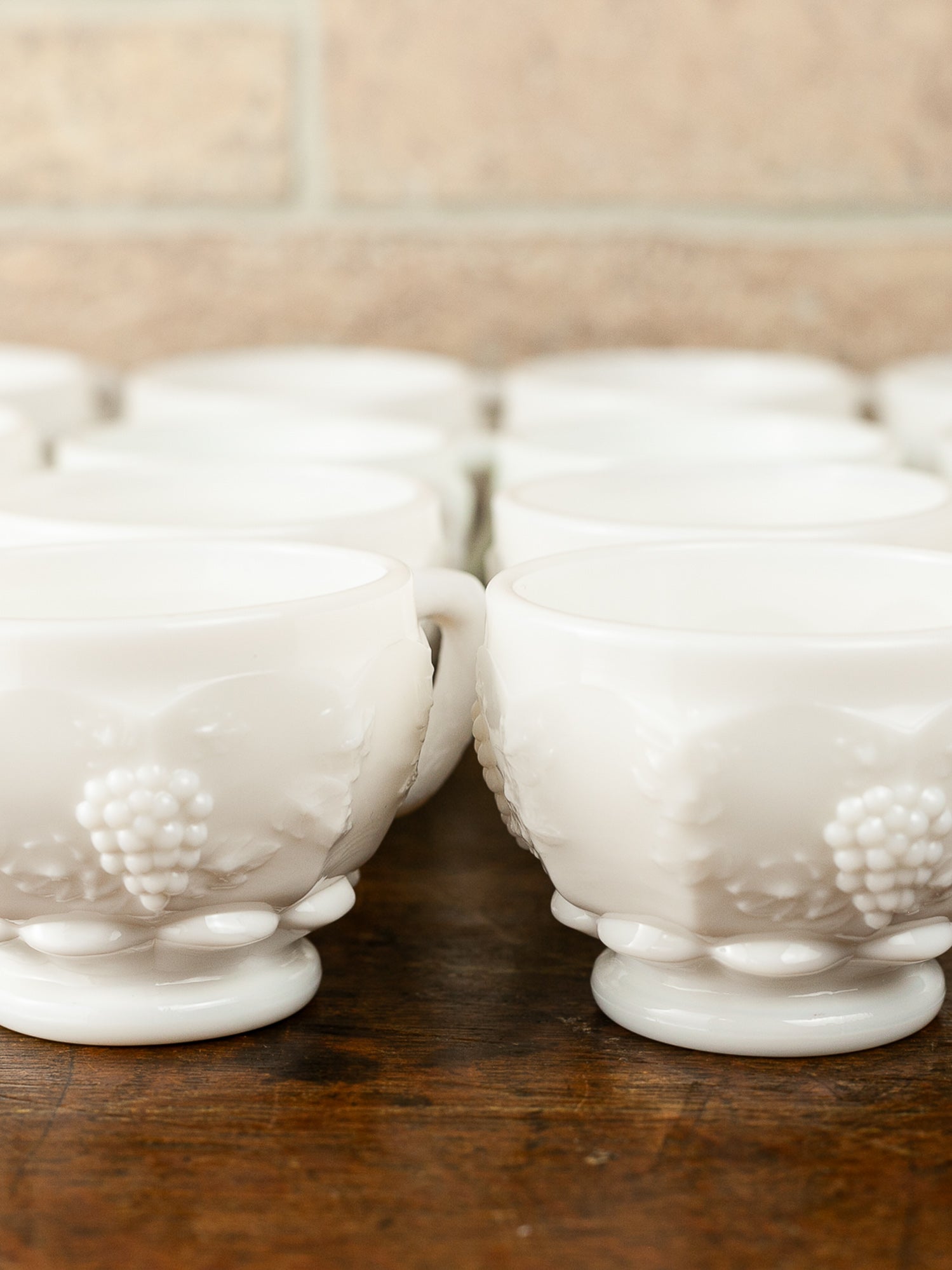 A set of white milk glass cups with embossed grapevine patterns and pedestal bases, arranged on a dark wooden surface with a blurred brick wall background.