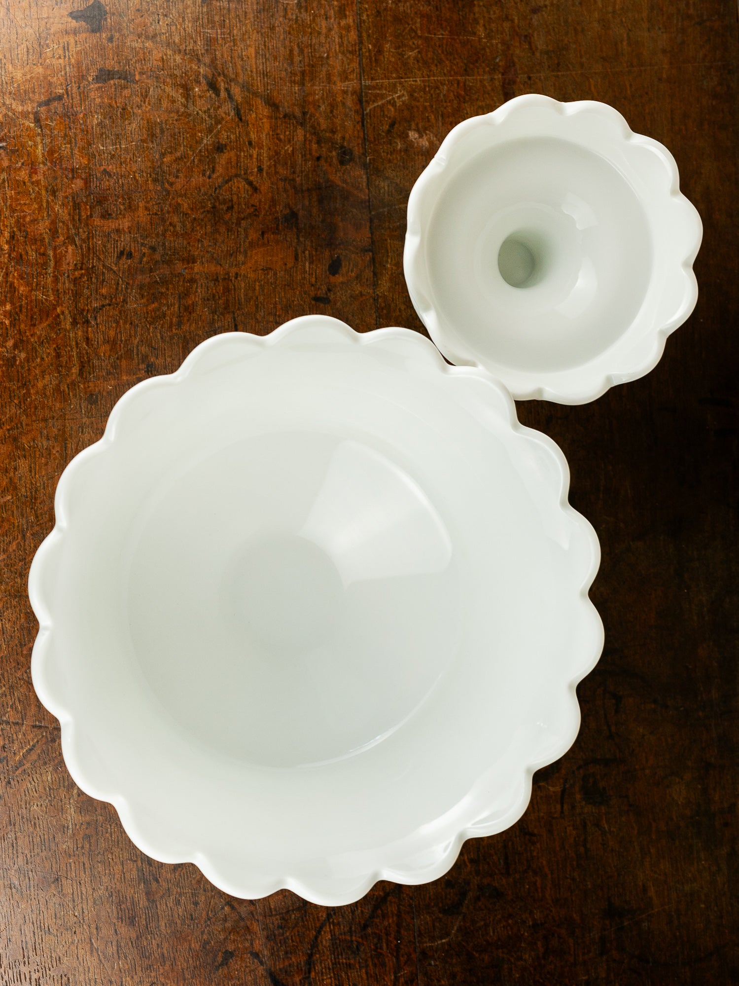 A white scalloped bowl and a matching small pedestal arranged on a dark brown wooden surface.