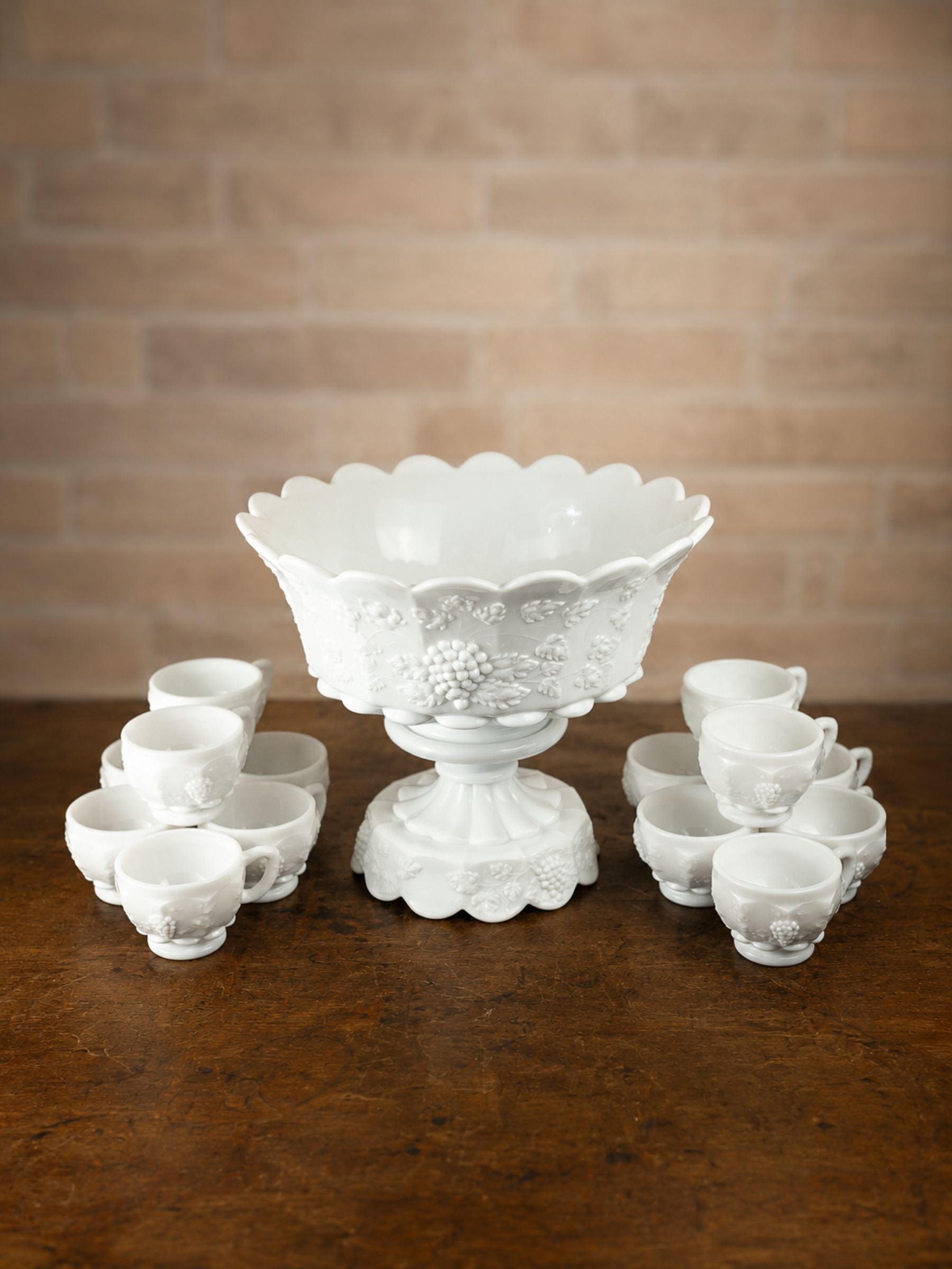 A white embossed glass punch bowl set with a pedestal base and multiple matching small cups, arranged on a dark wooden table against a warm brick wall background.