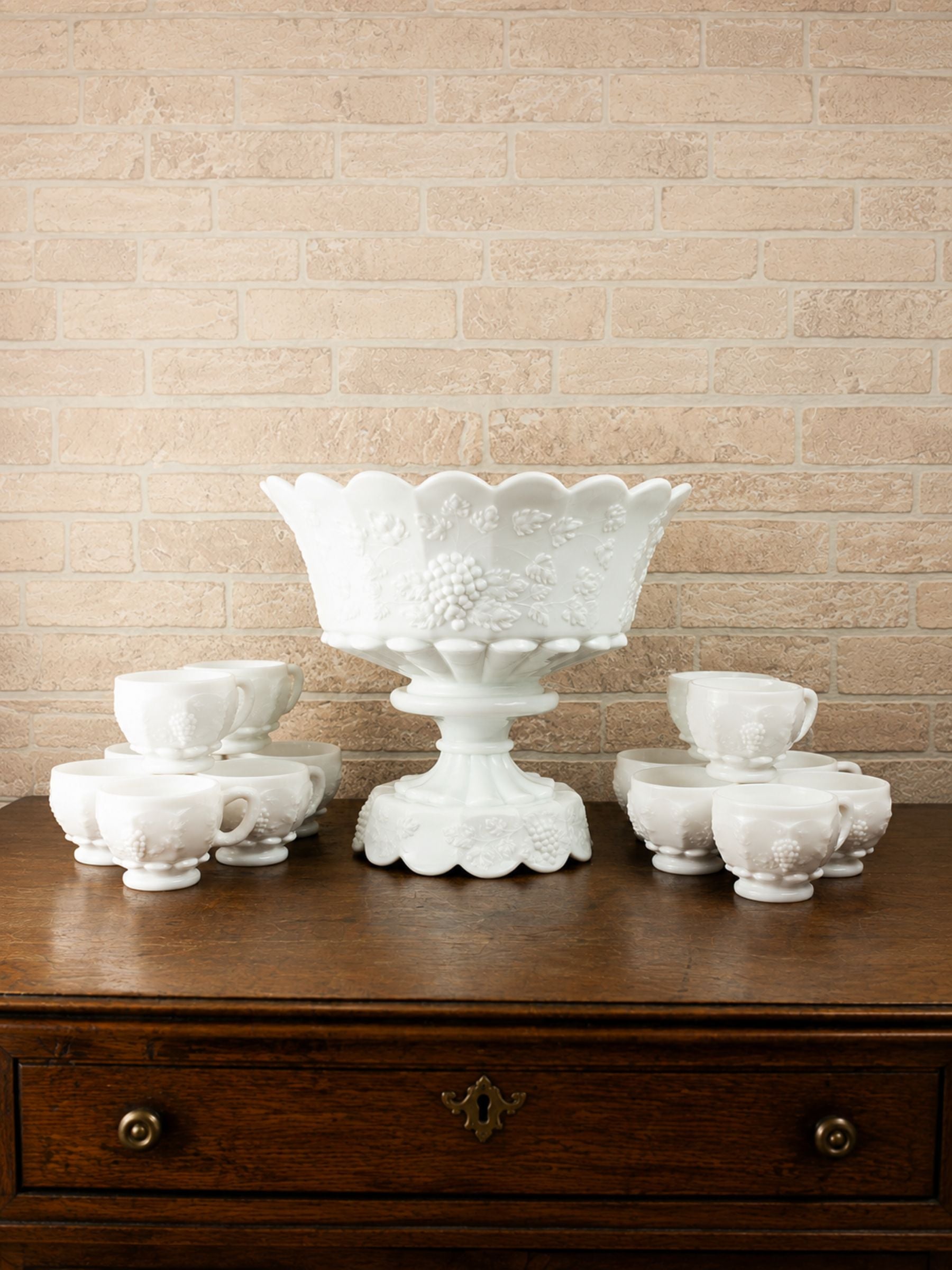 A white embossed glass punch bowl and matching cups arranged on a dark wooden chest against a beige brick wall.