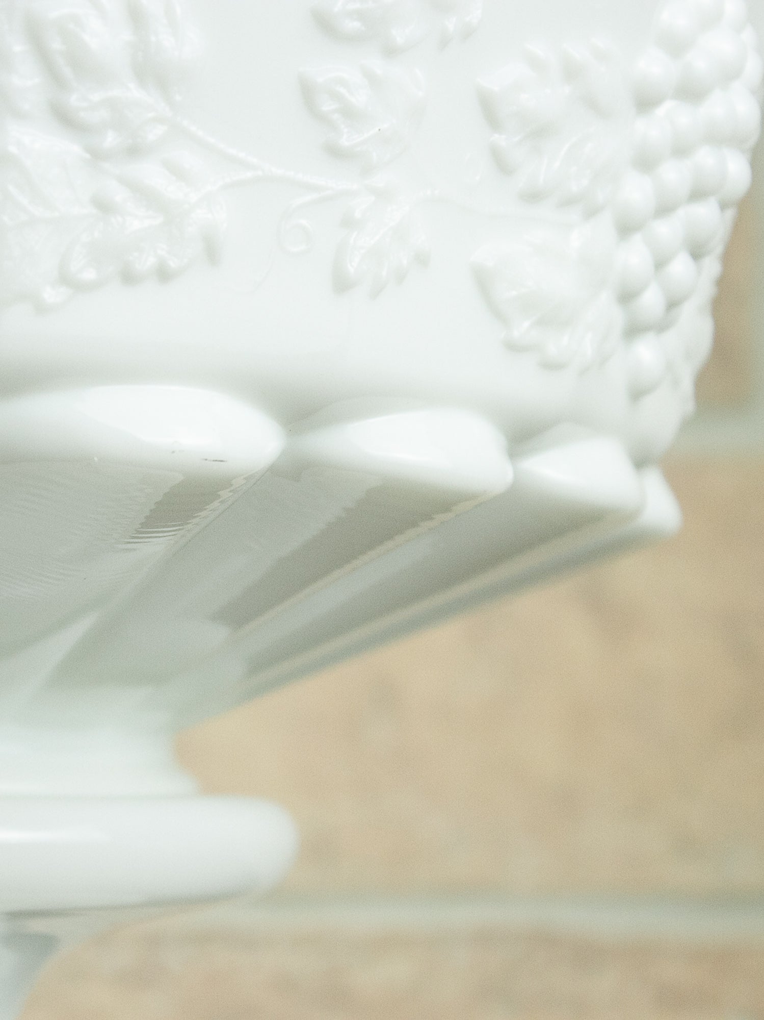 Close-up of a white embossed glass punch bowl with a floral and grapevine pattern, showing the textured side and scalloped rim against a soft beige background.