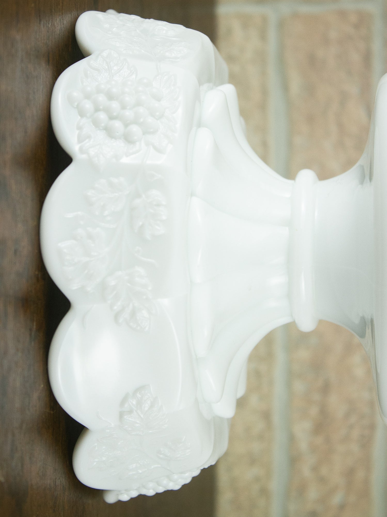 White milk glass pedestal base with embossed grapevine and fruit motifs, shown in a close-up side view against a dark wood and tiled background.