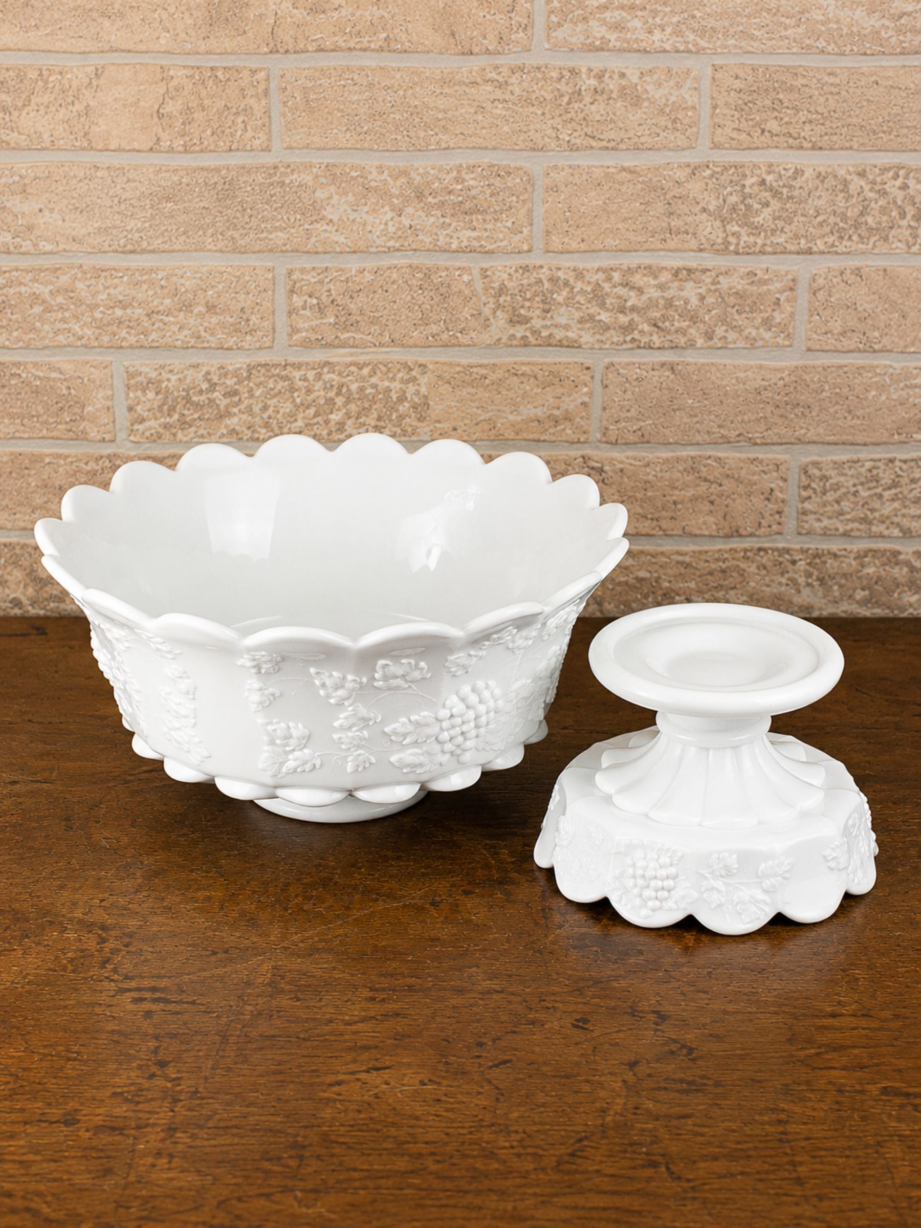 A white embossed glass bowl with a scalloped rim and matching small pedestal, displayed on a dark wood surface against a tan brick wall.