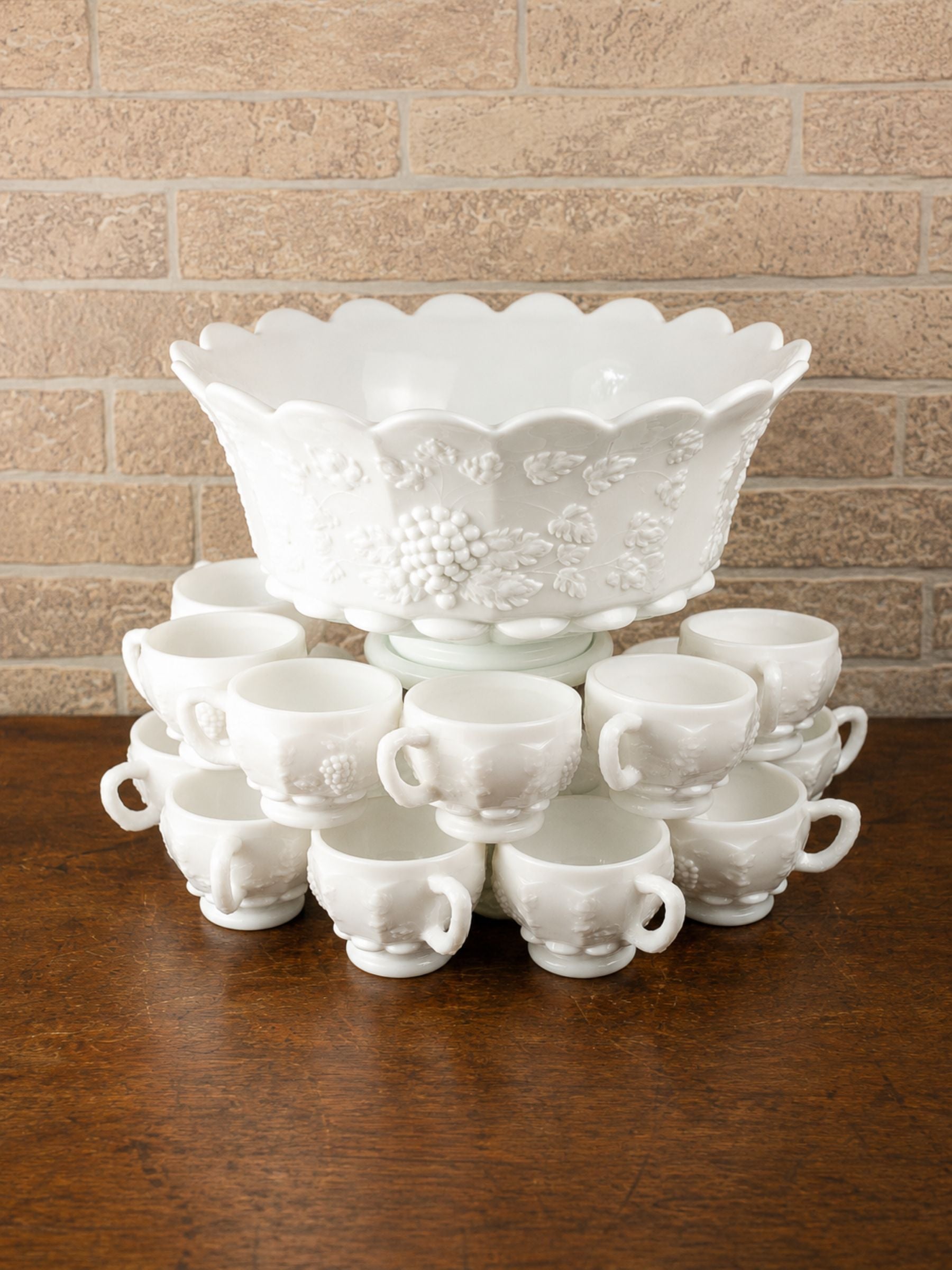 A white embossed glass punch bowl with a scalloped rim, displayed on a wooden table with fourteen matching small cups arranged underneath, against a tan brick wall background.