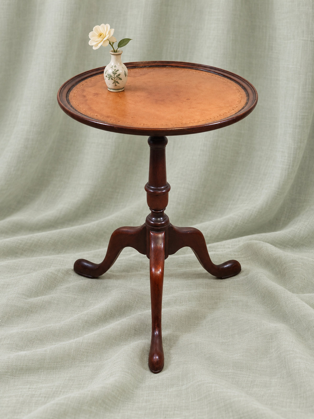Round Baker Furniture mahogany side table with a tan leather inset top, turned pedestal and tripod base, styled with a small flower vase.