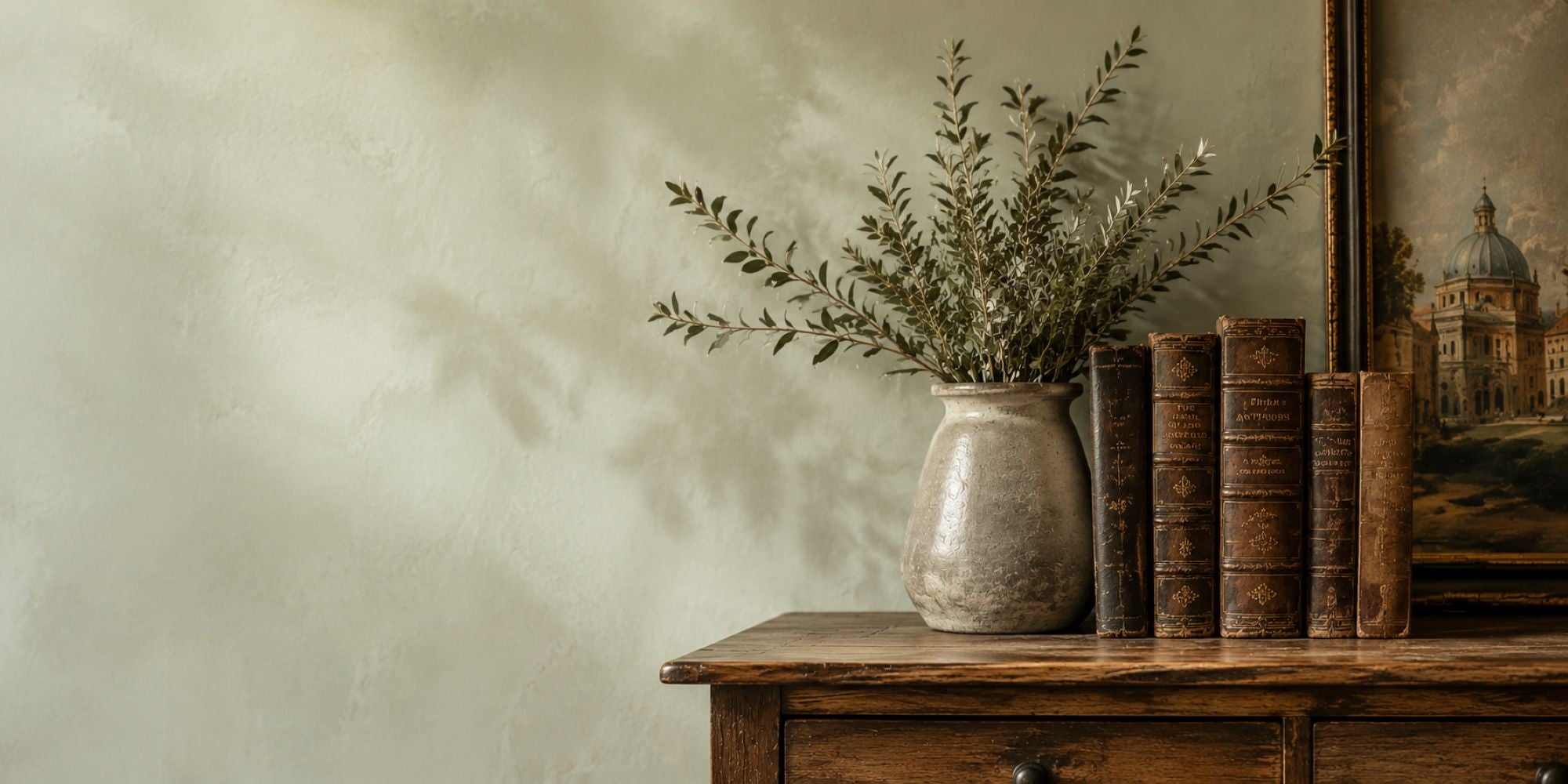 A rustic ceramic vase filled with dried greenery sits on a wooden cabinet beside a row of vintage brown books and a framed landscape painting against a textured beige wall.