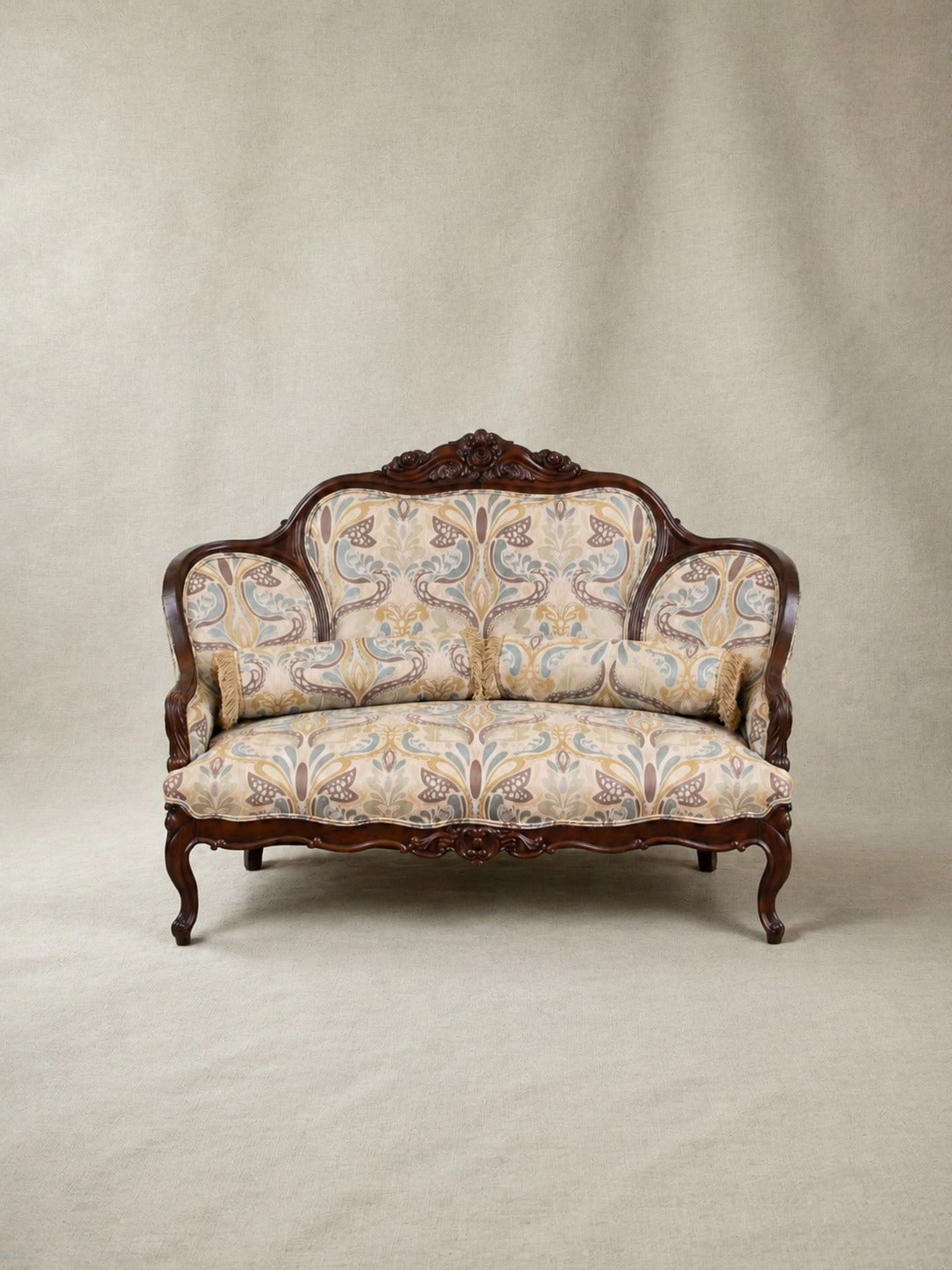 An antique-style loveseat with carved dark wood frames and patterned upholstery in beige, blue, and brown tones, photographed against a neutral beige backdrop.