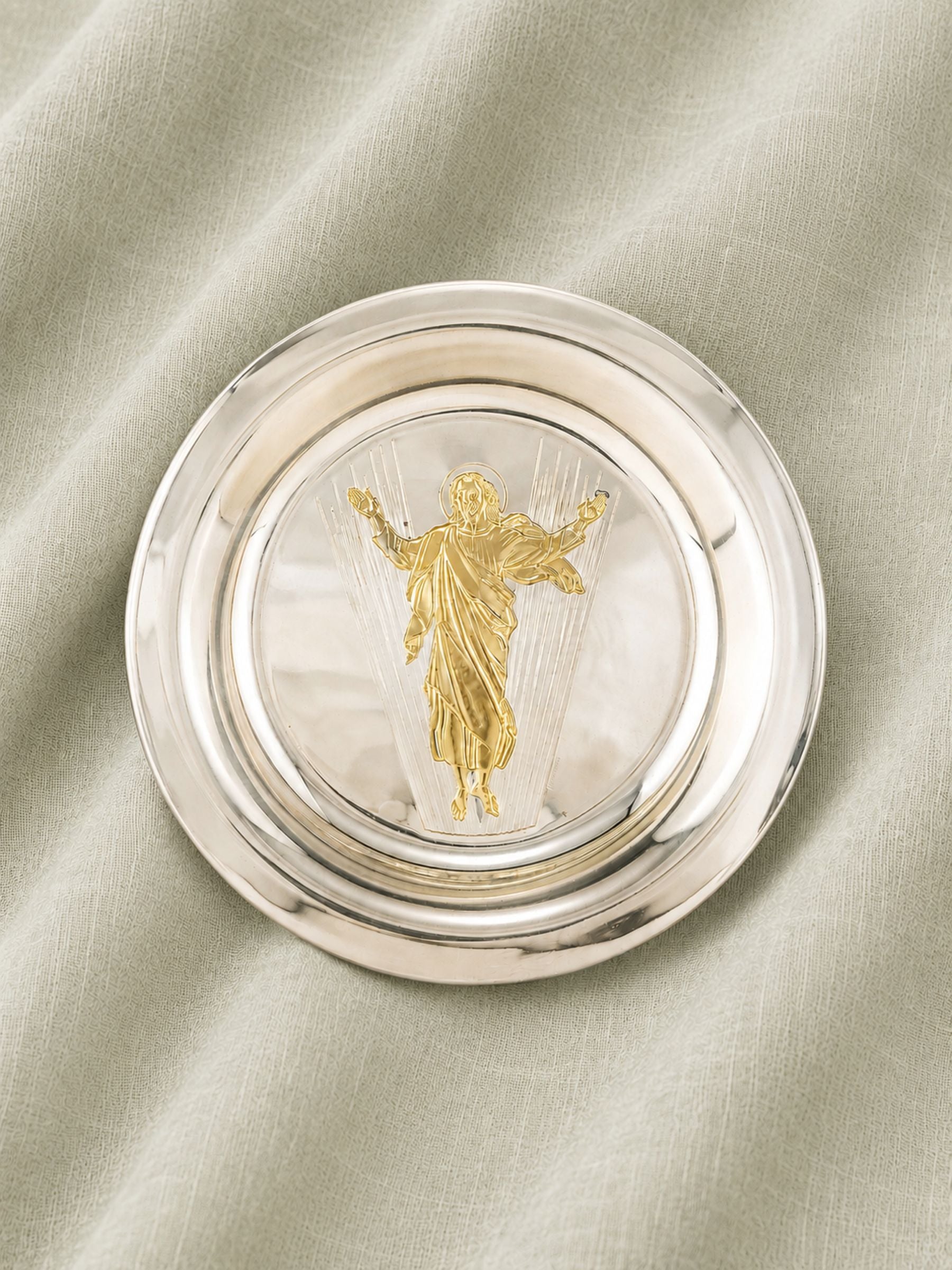 A round silver-toned metal collectors "resurrected Christ" plate with a gold-toned figure of Jesus Christ in the center, resting on a draped light beige fabric background.