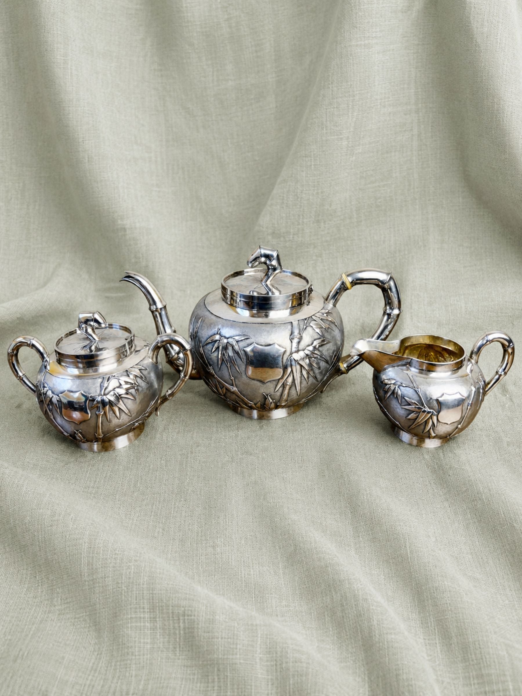 A three-piece Sterling silver teapot with milk and sugar bowl set with embossed bamboo designs, displayed on a rustic wooden table against a light brick wall background.