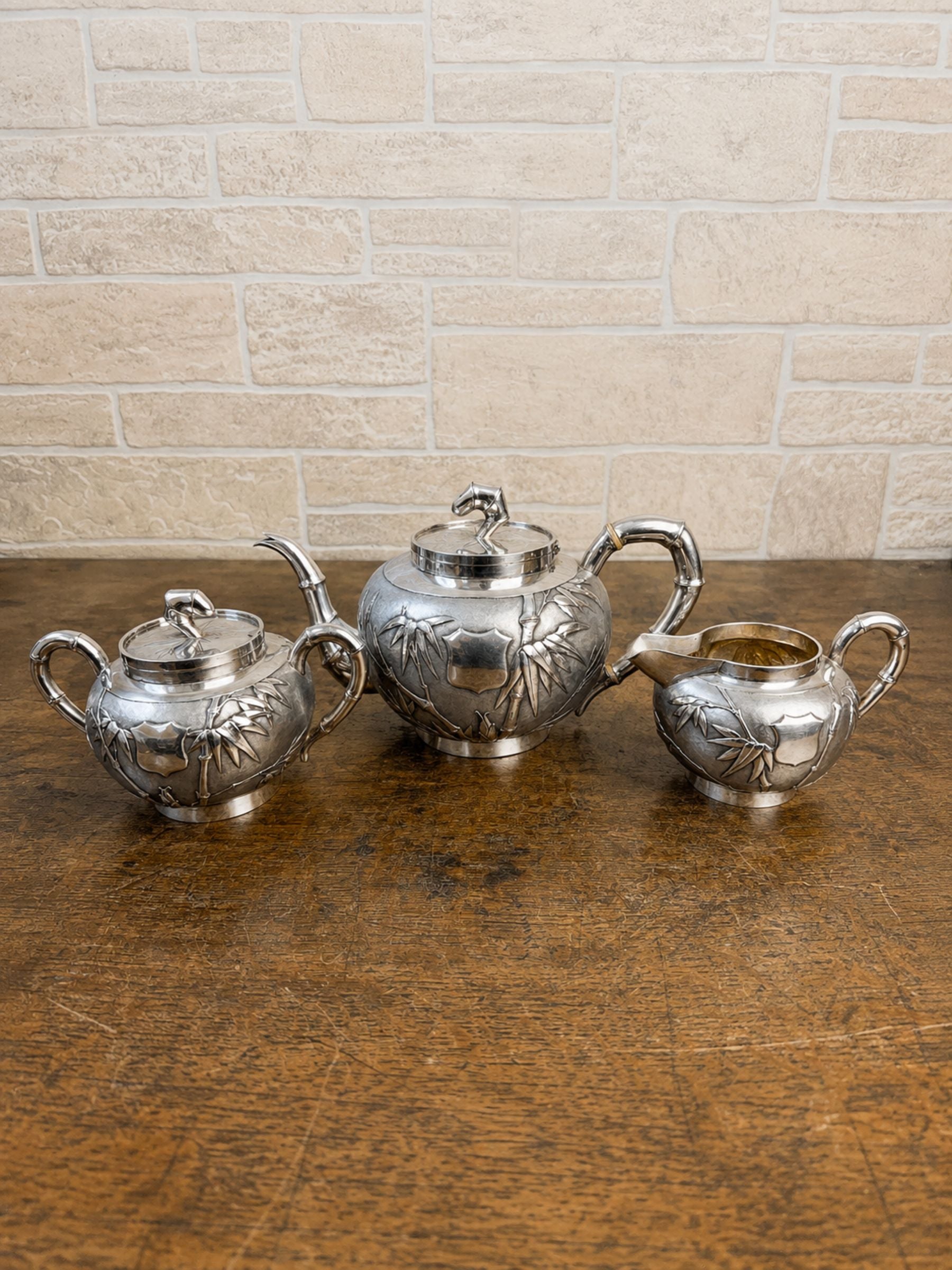 A three-piece silver teapot and sugar bowl set with embossed bamboo designs, displayed on a rustic wooden table against a light brick wall background.