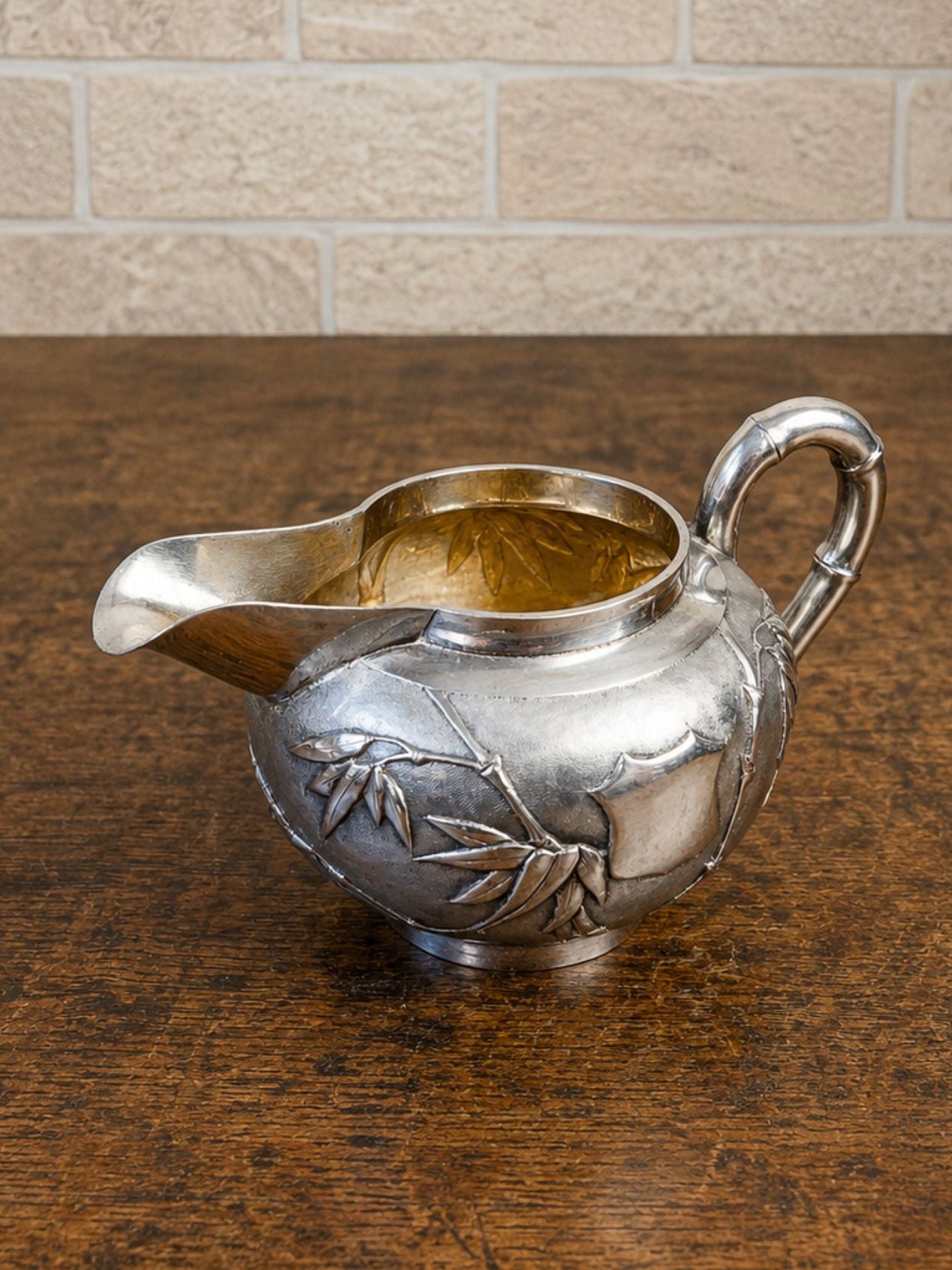 A vintage sterling silver creamer or sauceboat with embossed leaf detailing, resting on a dark wooden table against a light beige brick wall.