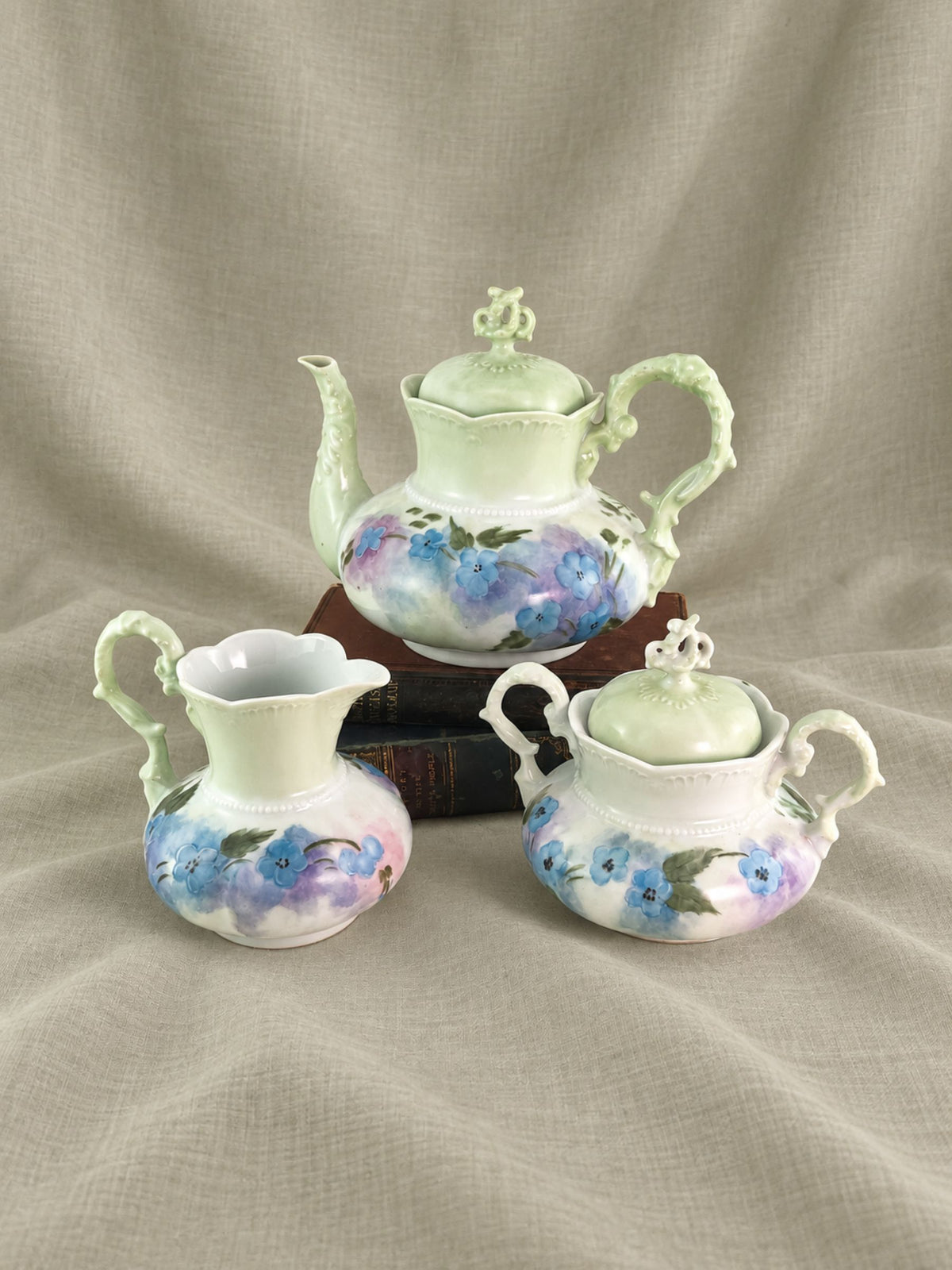 A three-piece ceramic tea and coffee set with a teapot, creamer, and sugar bowl, decorated with blue and purple floral patterns on a white base, set against a draped beige fabric background.