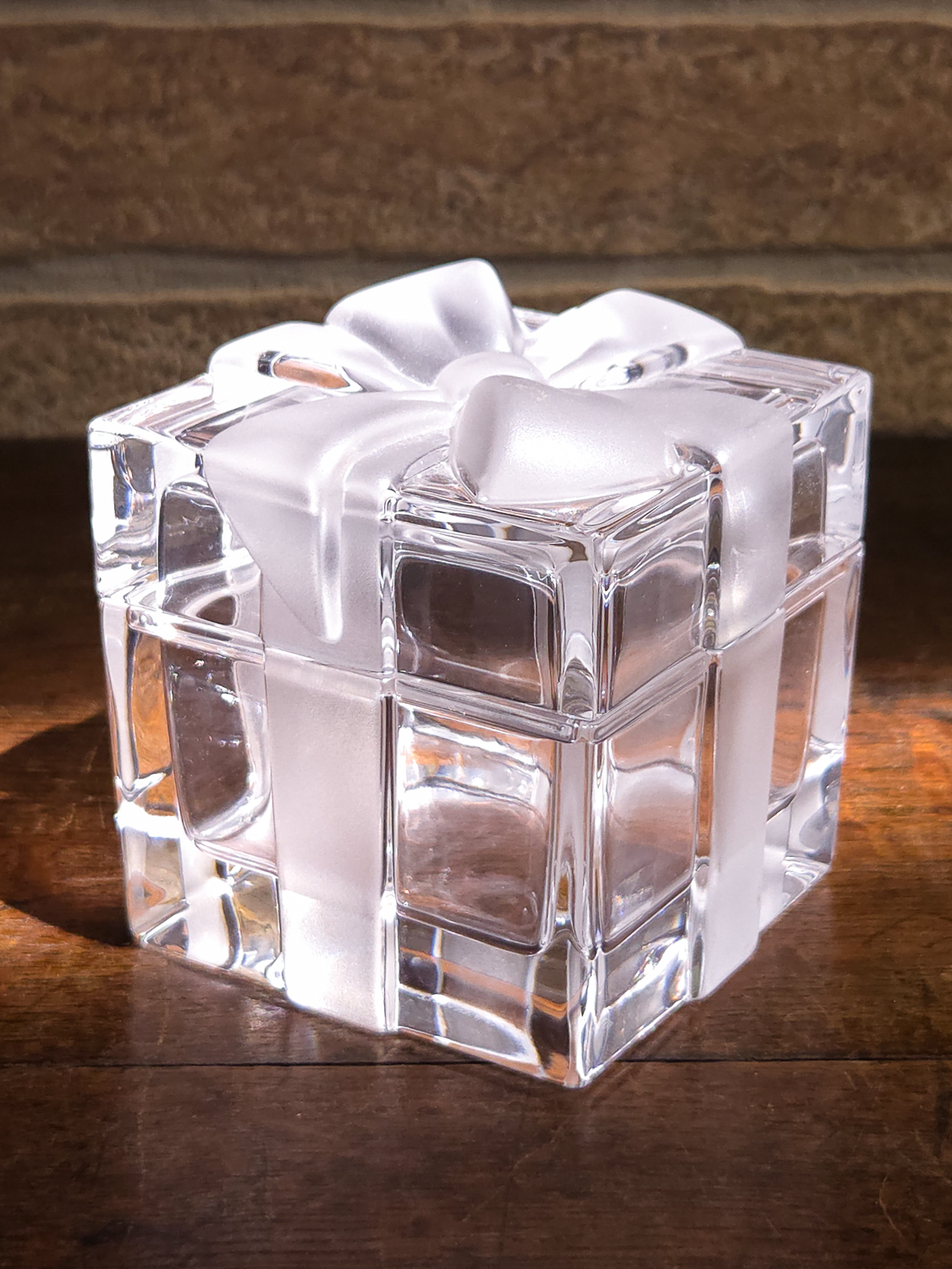 A clear glass gift box with a frosted white satin ribbon and bow, placed on a dark wooden surface with a stone wall background.