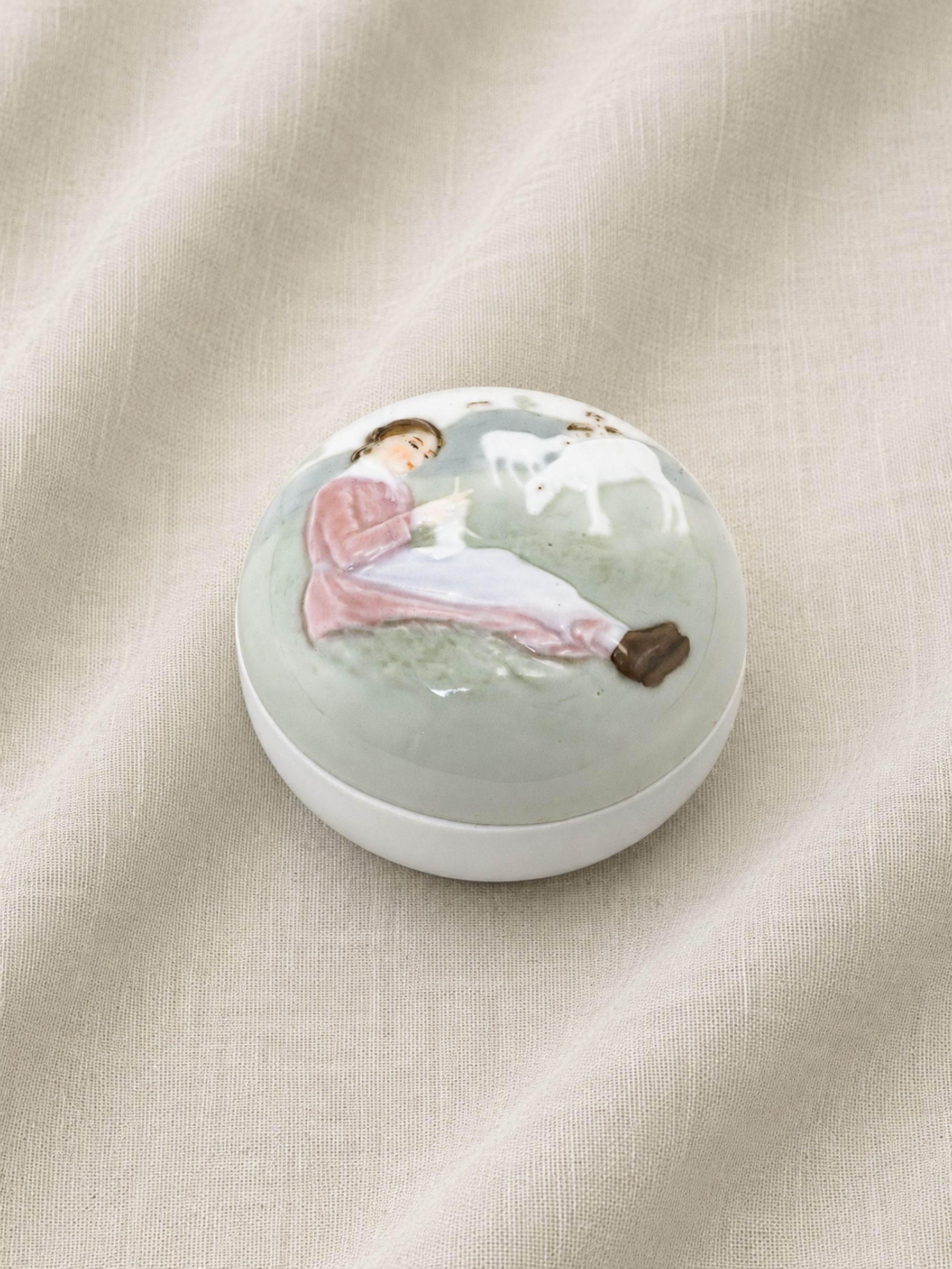 A round antique porcelain trinket or ring box by Karl Ens of Volkstedt, Germany,  with a glossy lid featuring a hand-painted illustration of a girl in a pink dress sitting on the ground next to a white sheep, set against a draped beige fabric background.