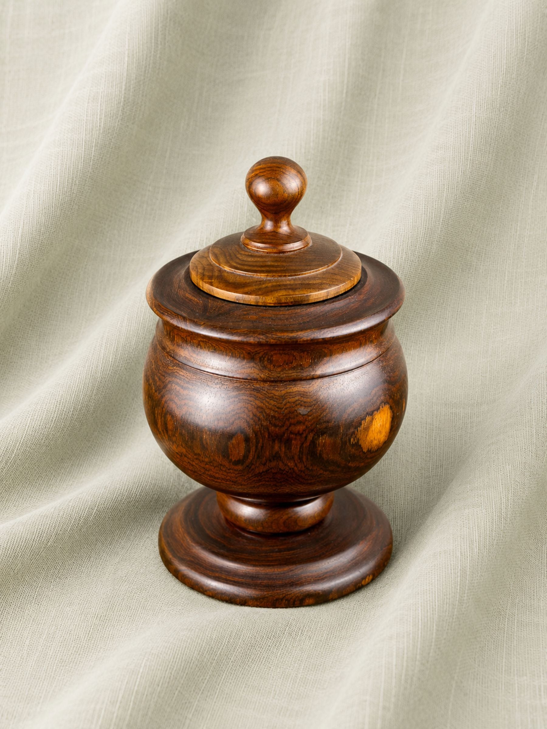 A round wooden lidded container or jar with a turned wooden knob on top, photographed against draped light beige fabric.