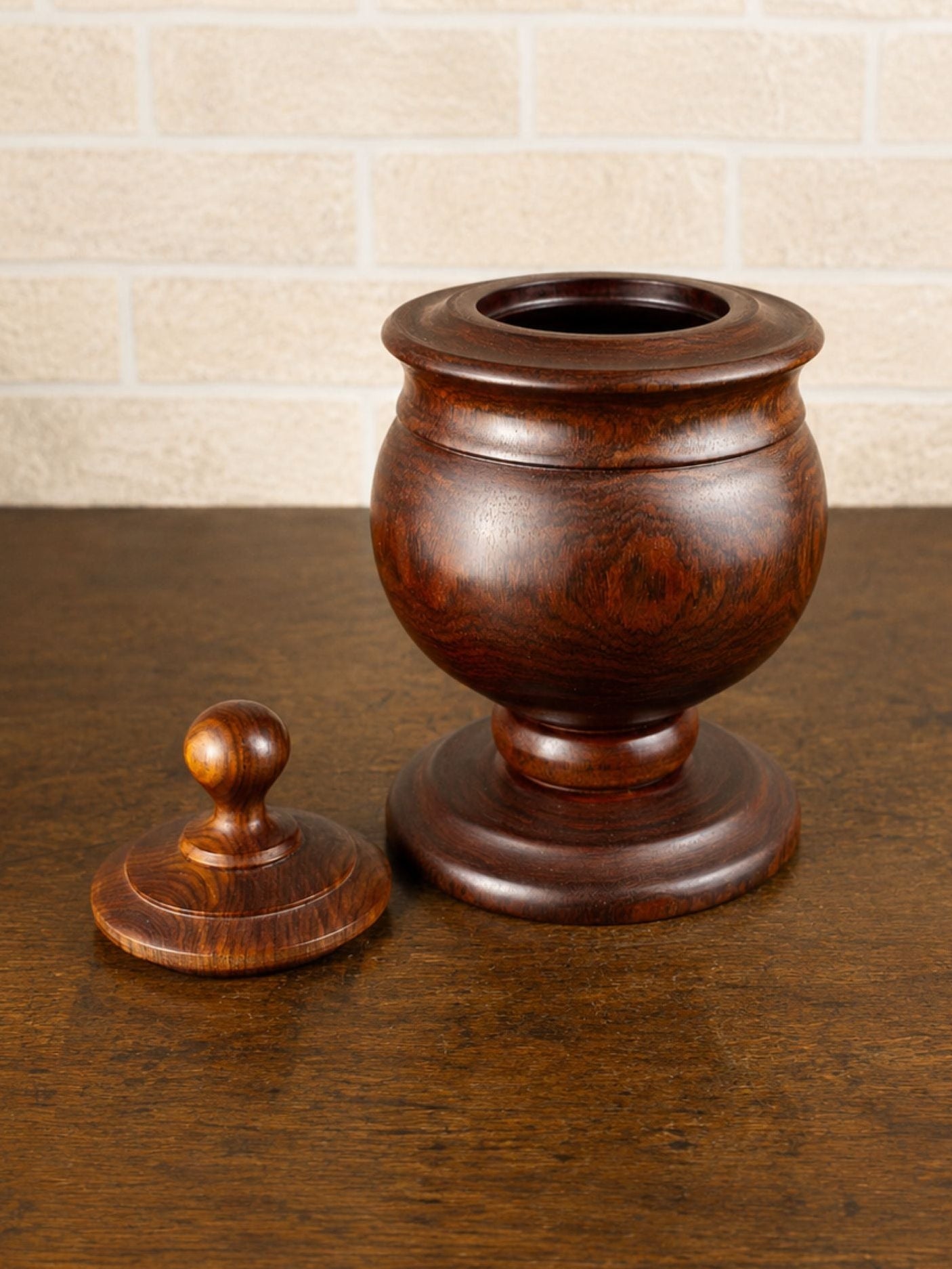 A dark brown wooden urn-shaped container with a matching lid placed beside it on a wooden tabletop, set against a light beige brick wall background.