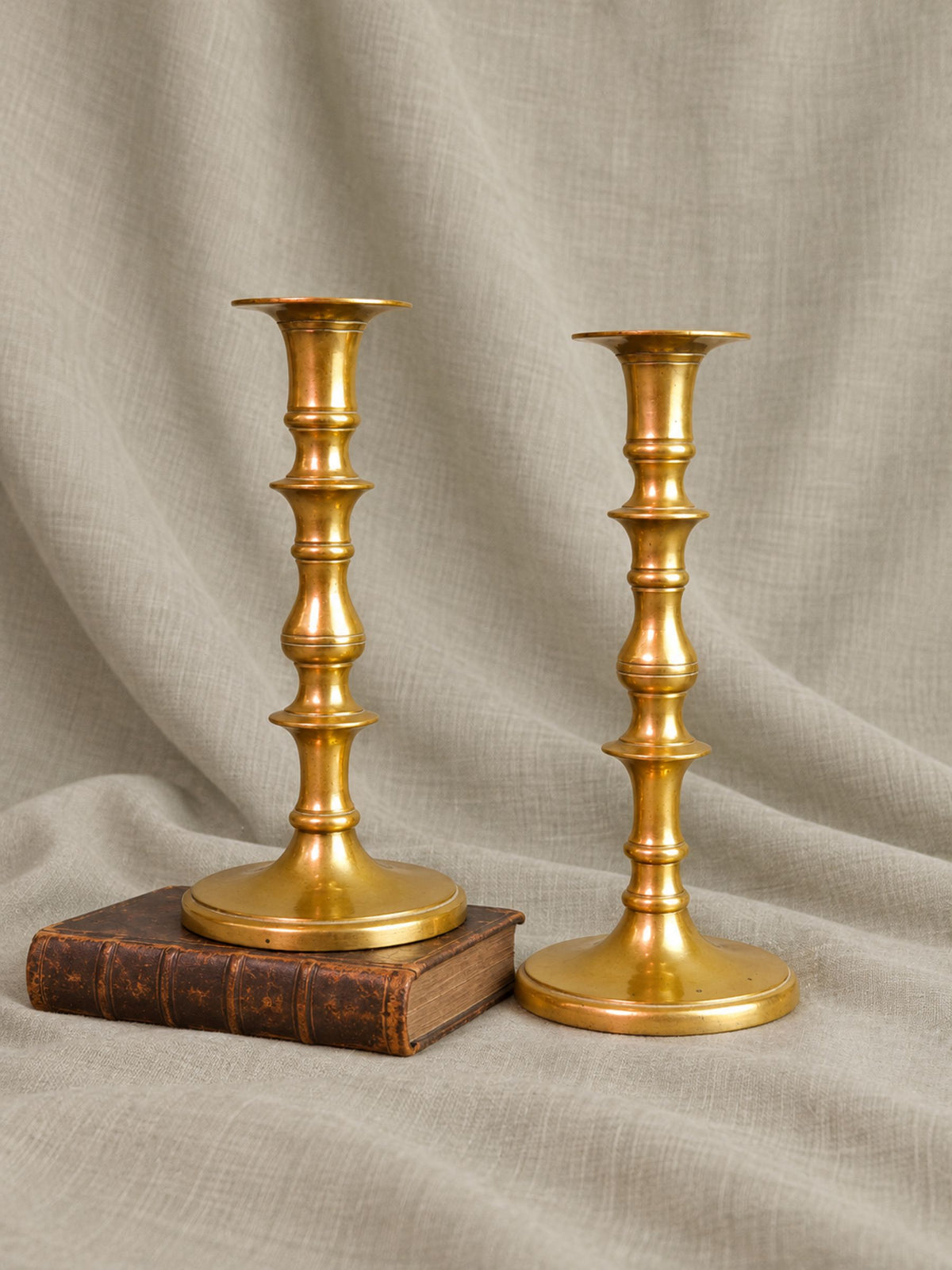 A pair of Victorian brass candlesticks with a classic turned-bead design, displayed on a draped beige linen fabric backdrop. One candlestick stands on a vintage brown leather-bound book, while the other stands upright beside it.