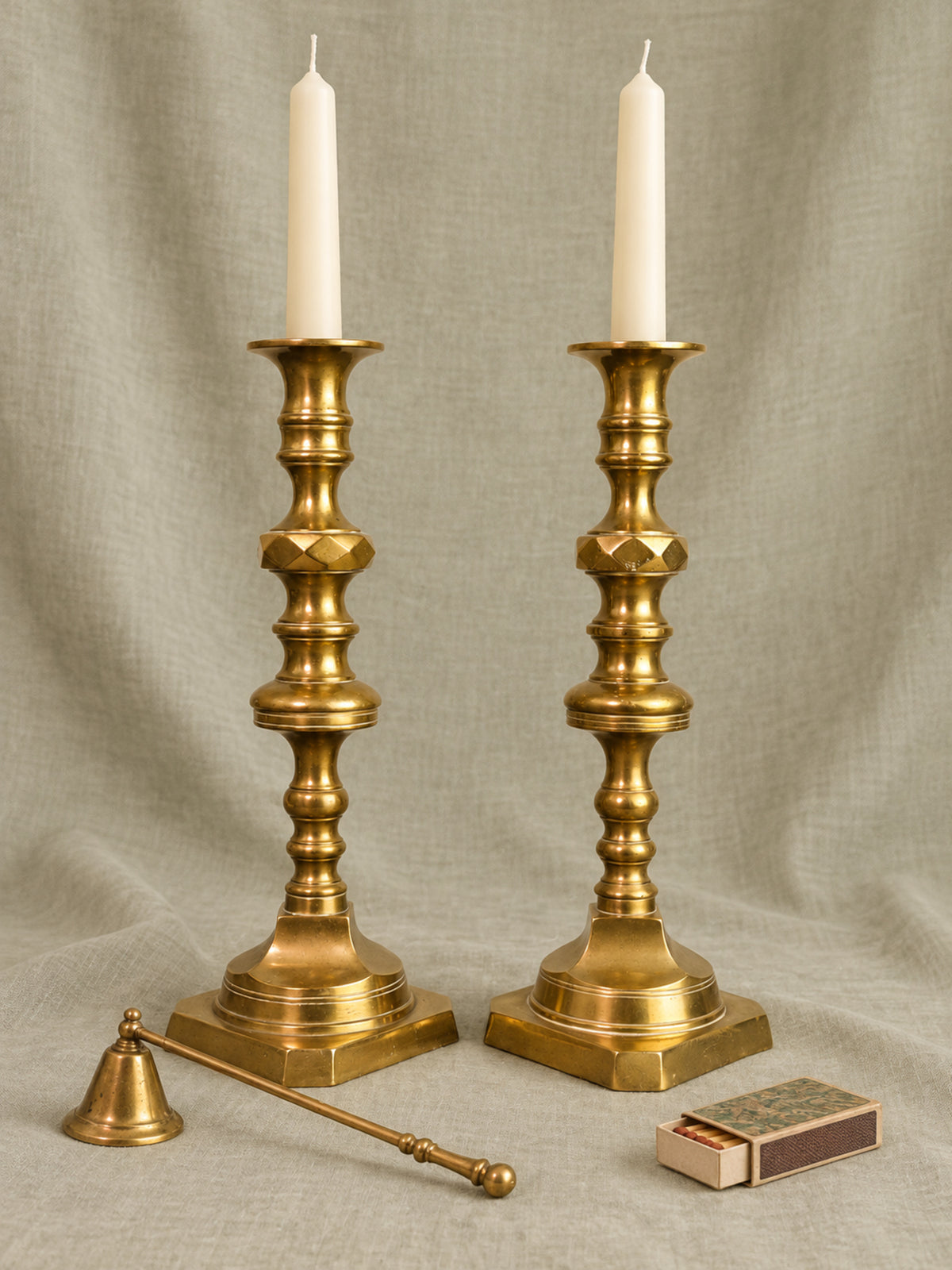 A pair of ornate brass candlestick holders with white taper candles, displayed on a draped beige fabric background with a brass bell and a matchbox as props.