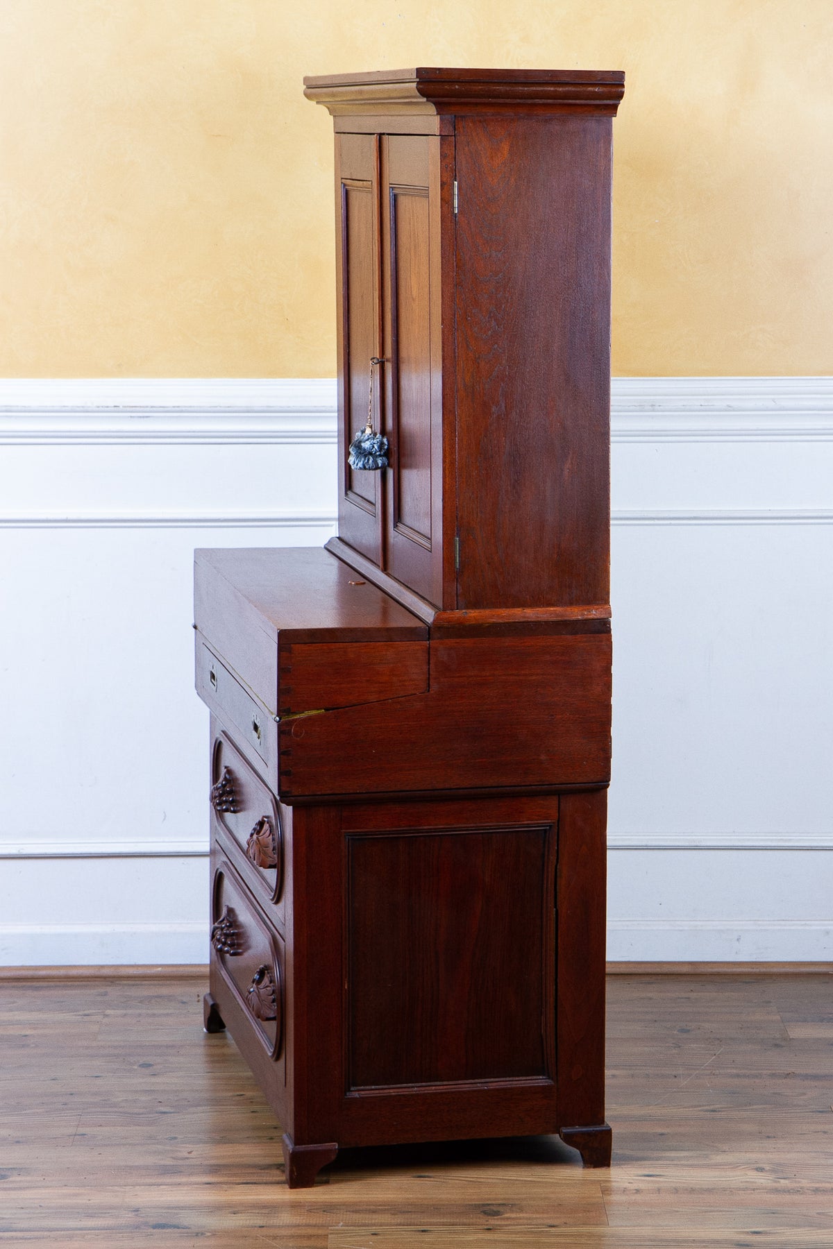 A dark reddish-brown wooden writing desk with an attached hutch cabinet, shown in a home interior setting with a yellow and white paneled wall and wood floor.