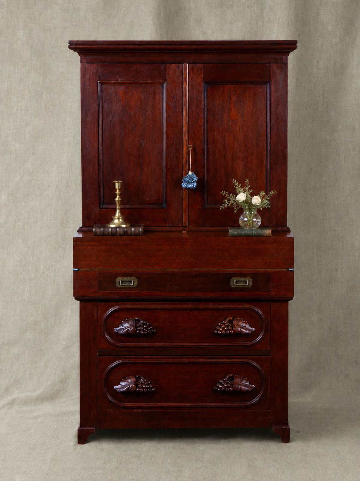 A dark reddish-brown wooden secretary desk with two upper doors, a middle drawer, and two lower drawers with decorative carved handles, photographed against a neutral beige fabric backdrop.
