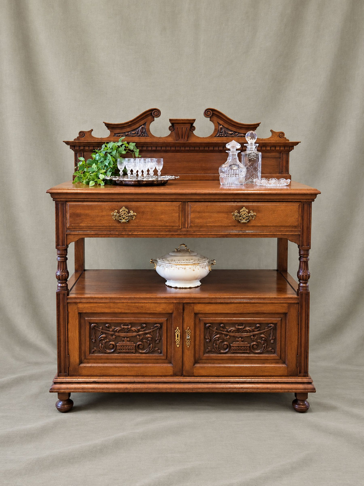 Antique carved wood Victorian cabinet with brass hardware, glass decanters, stemware, greenery, and a covered tureen.