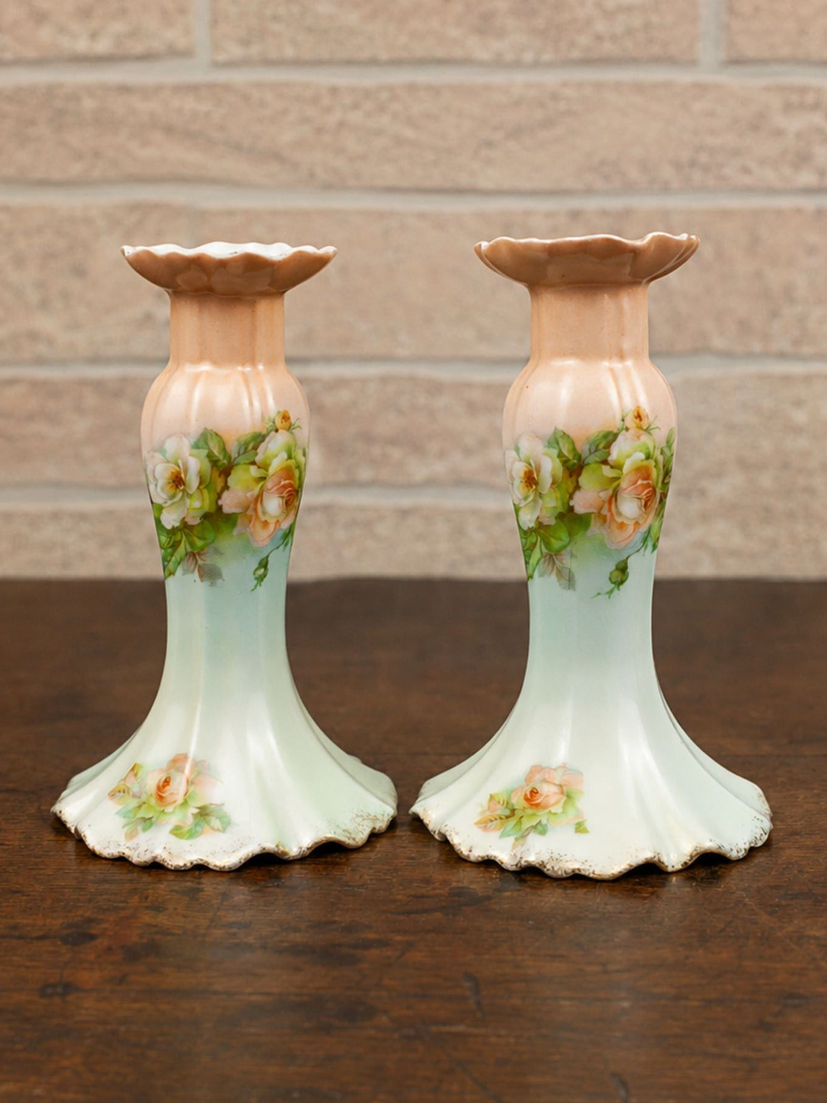 A pair of vintage ceramic candlesticks with a peach-colored upper section, a pale green flared base, and a hand-painted floral design in the center, displayed on a dark wooden table against a light brick wall.
