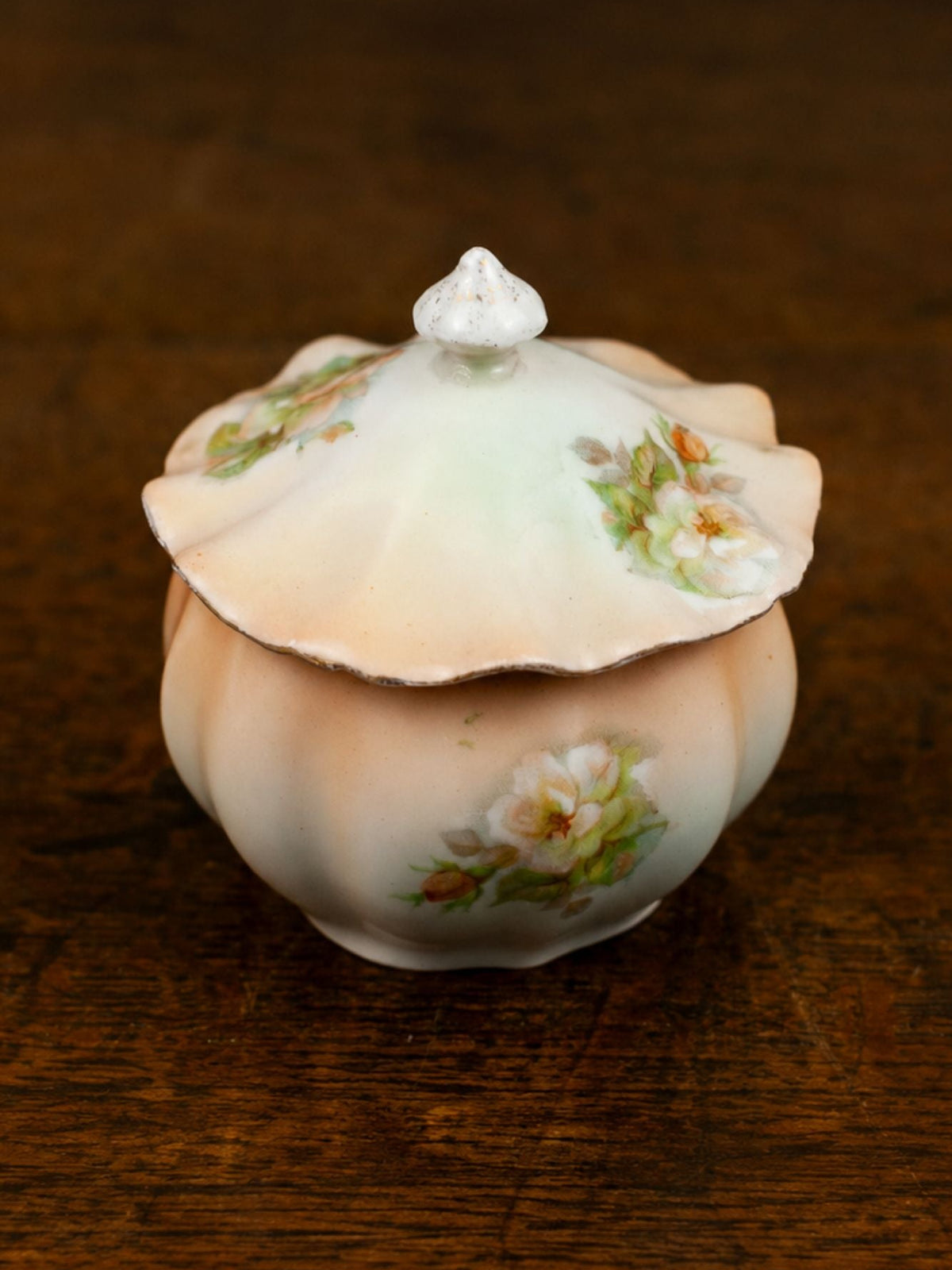 A vintage ceramic lidded sugar bowl with a scalloped lid and delicate floral hand-painted details, resting on a dark wooden surface.