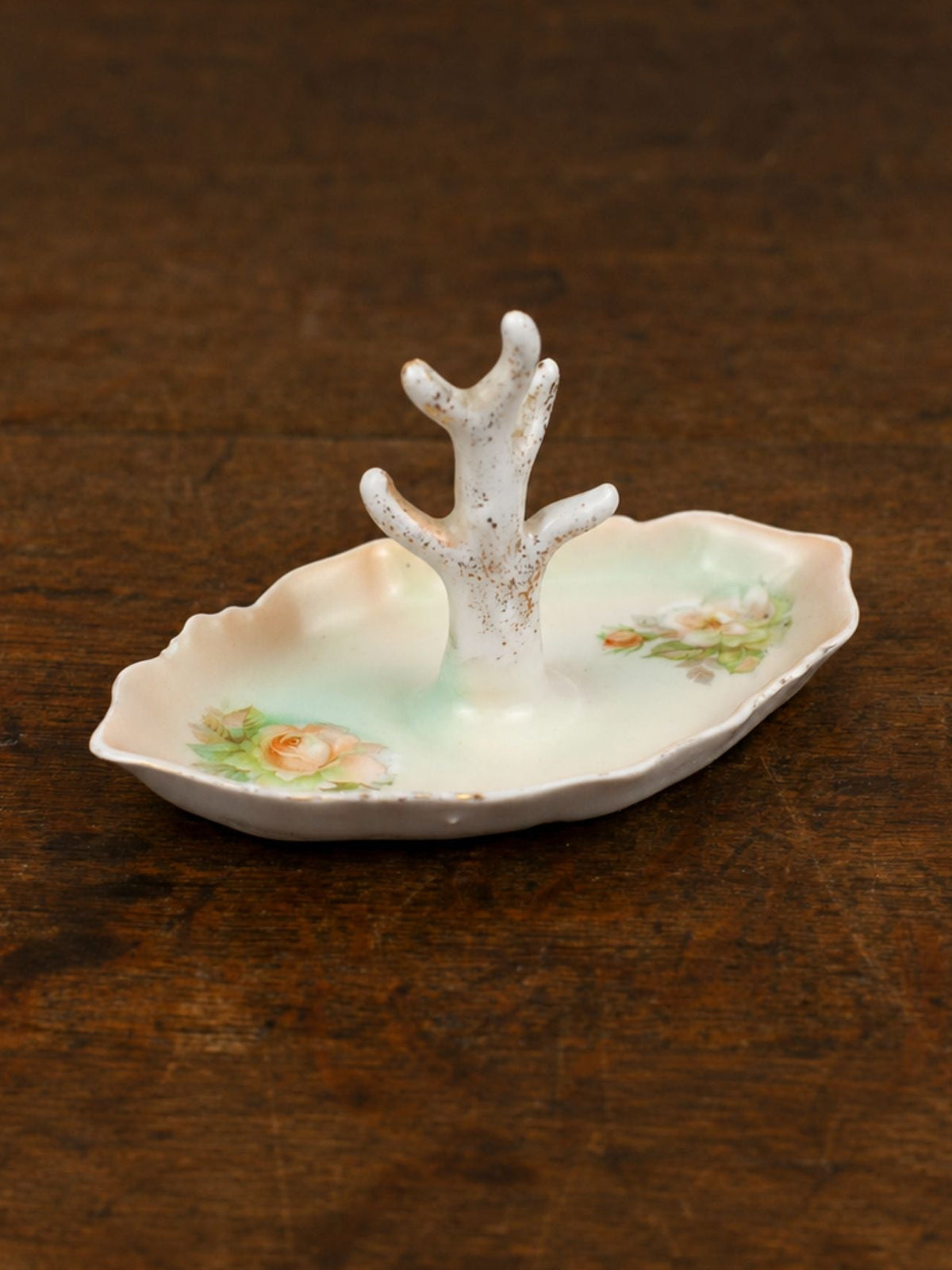 A small decorative jewelry dish with a tree-like center, featuring a pale green and white glaze with delicate floral details, set on a dark brown wooden surface.