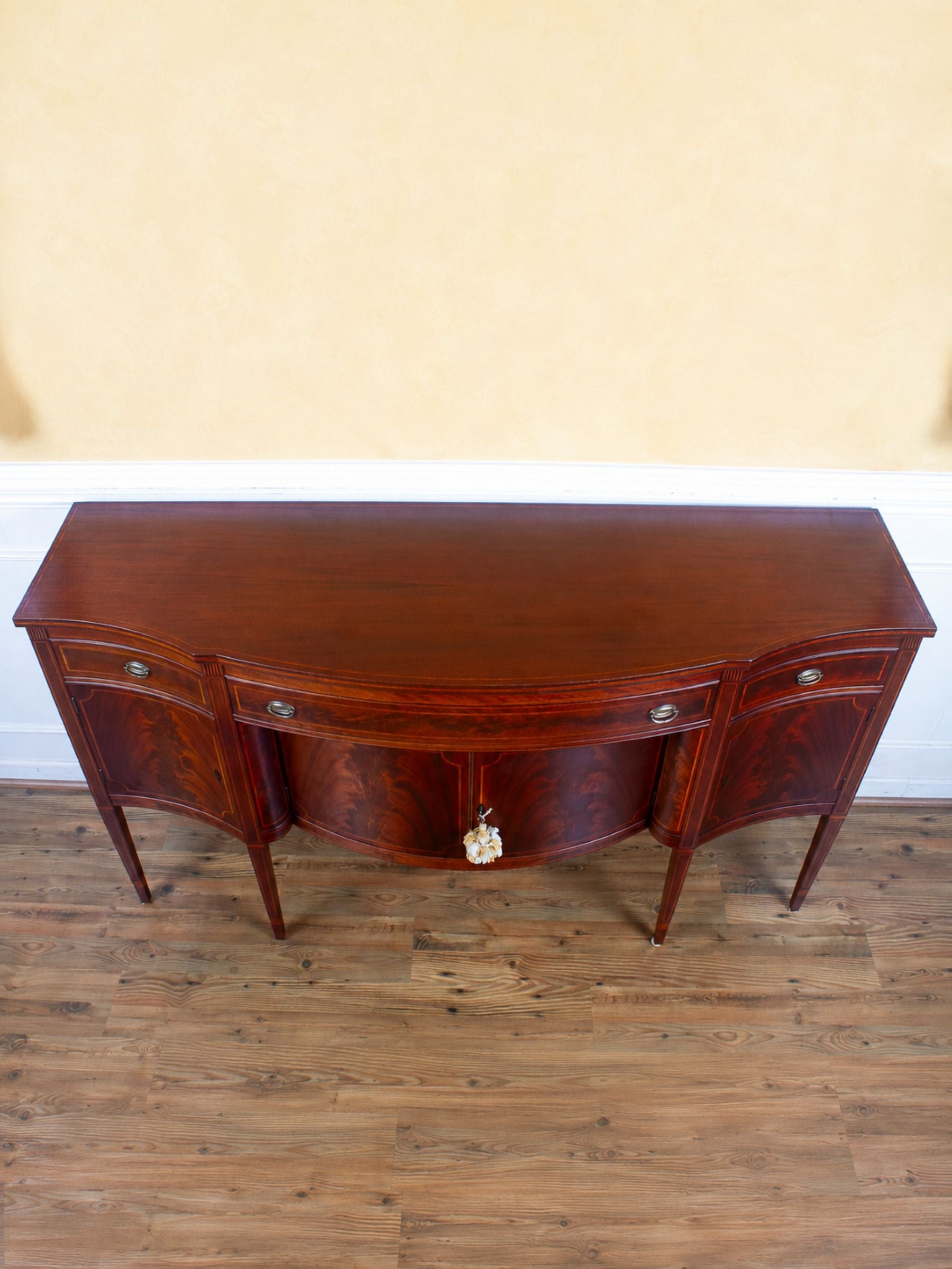 A polished reddish-brown wooden sideboard with curved front panels, three drawers, and small round metal knobs, shown against a beige wall and wood floor. 