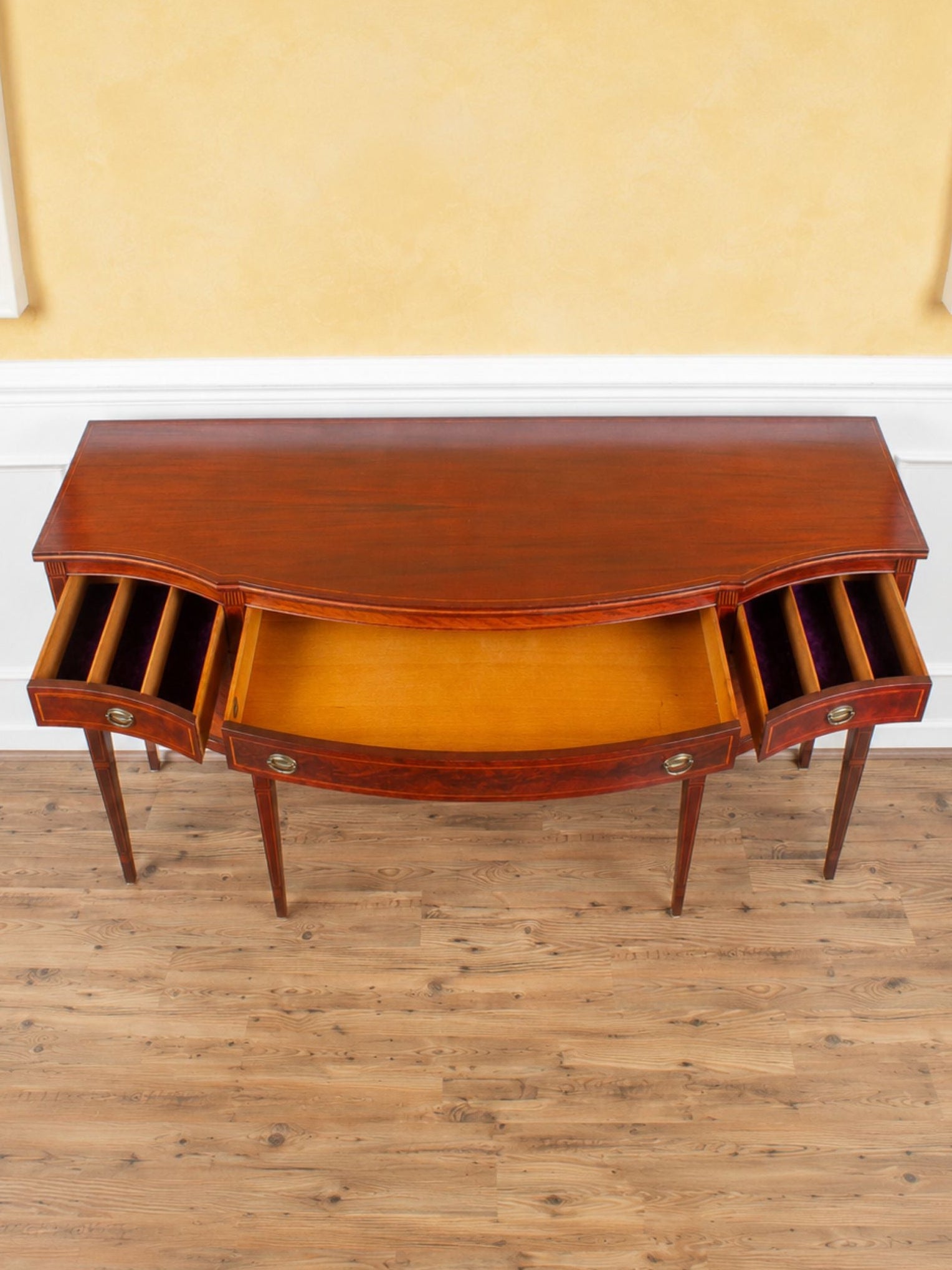 A curved mahogany sideboard with open drawers and a warm orange-brown interior, set against a yellow wall and light wood floor.