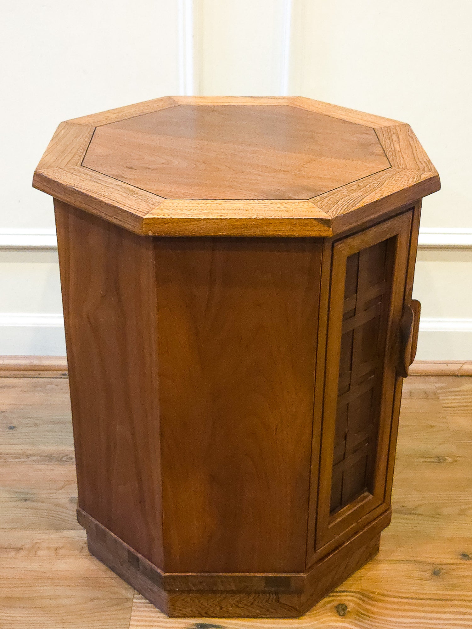 A hexagonal wooden side table or cabinet with a warm medium-brown finish, featuring a slanted octagonal top and a glass-paneled door on the right side, set on a wooden floor against a white wall.