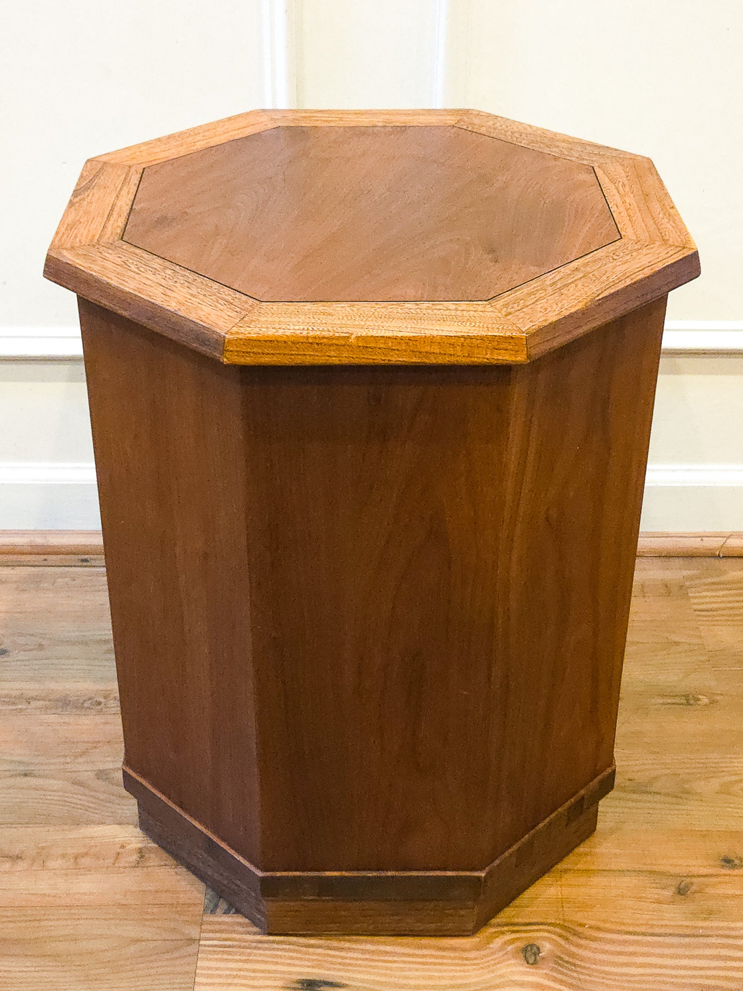 A hexagonal wooden side table or end table with a warm medium-brown finish and a lighter wood octagonal border around the top, shown on a wood floor against a white wall.