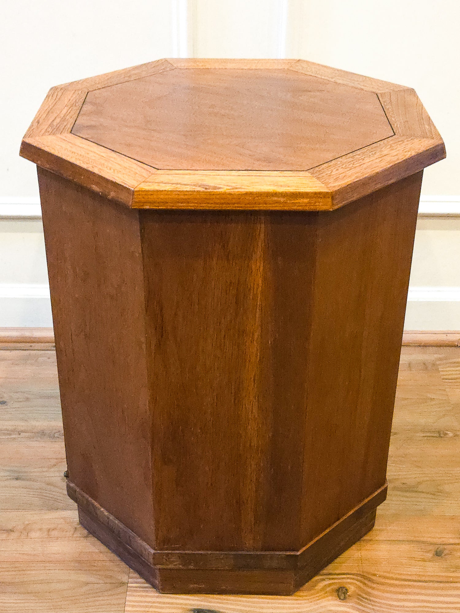 A hexagonal wooden side table or end table with a warm medium-brown finish, set on a light wood floor against a white wall.