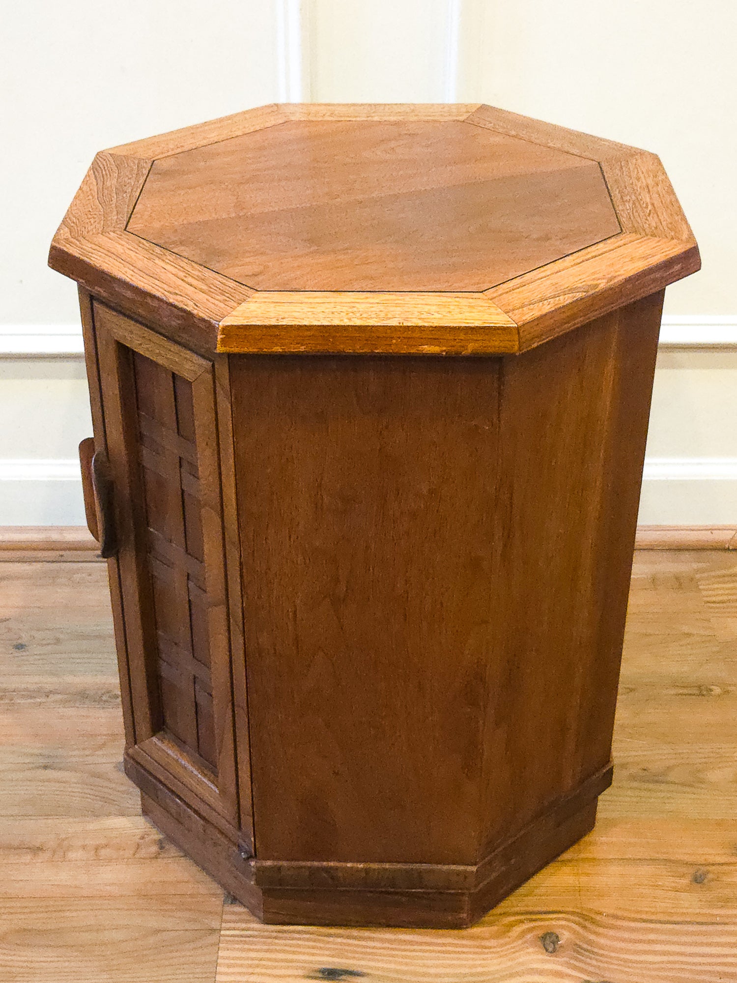 A hexagonal wooden side table or cabinet with a warm medium-brown finish, featuring a paneled front door and octagonal top, set on a wooden floor against a white wall.