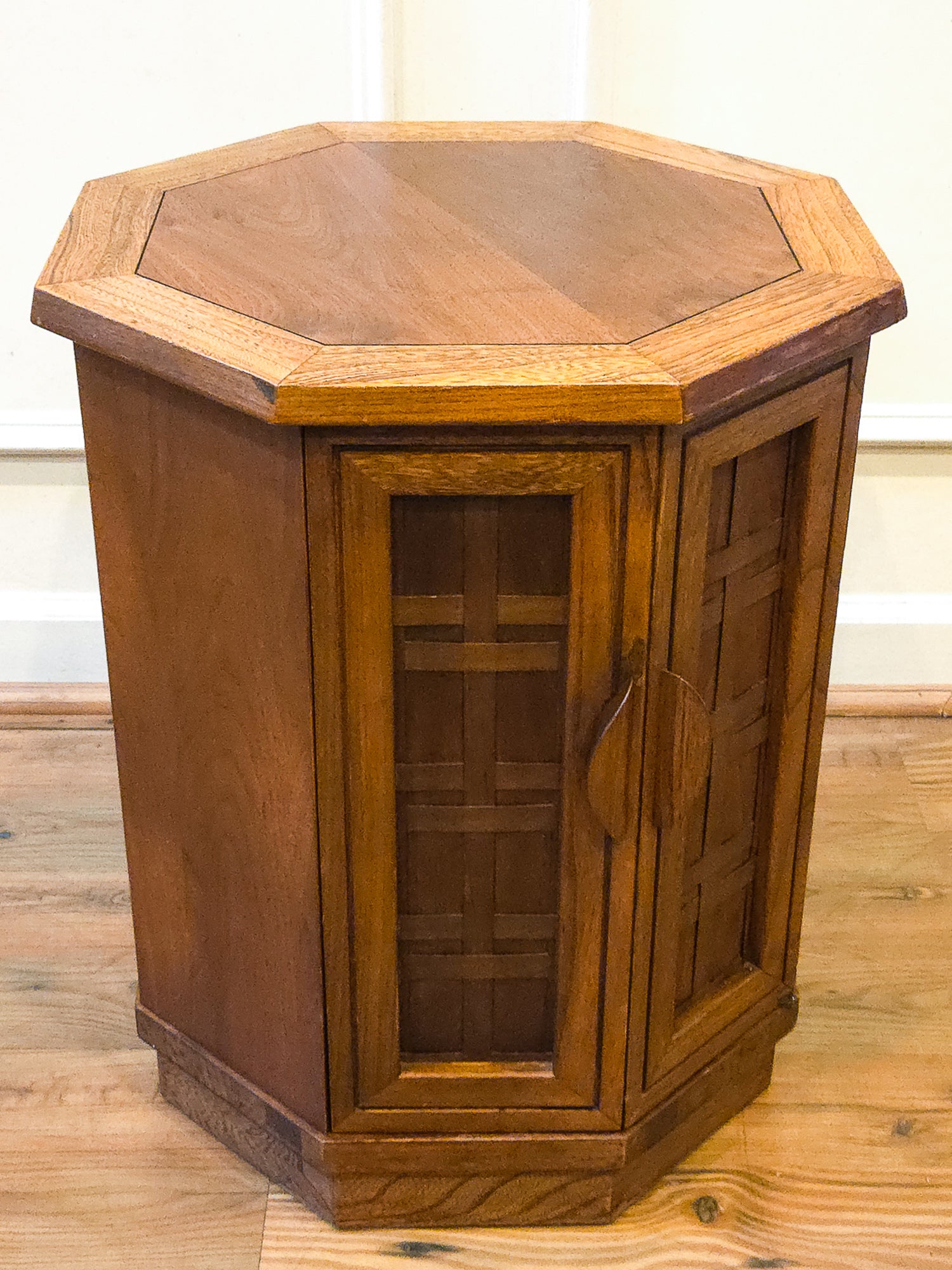 A hexagonal wooden cabinet end table with a warm medium-brown finish, featuring two paneled doors with geometric lattice patterns and a single central handle, set on a wooden floor against a white wall.