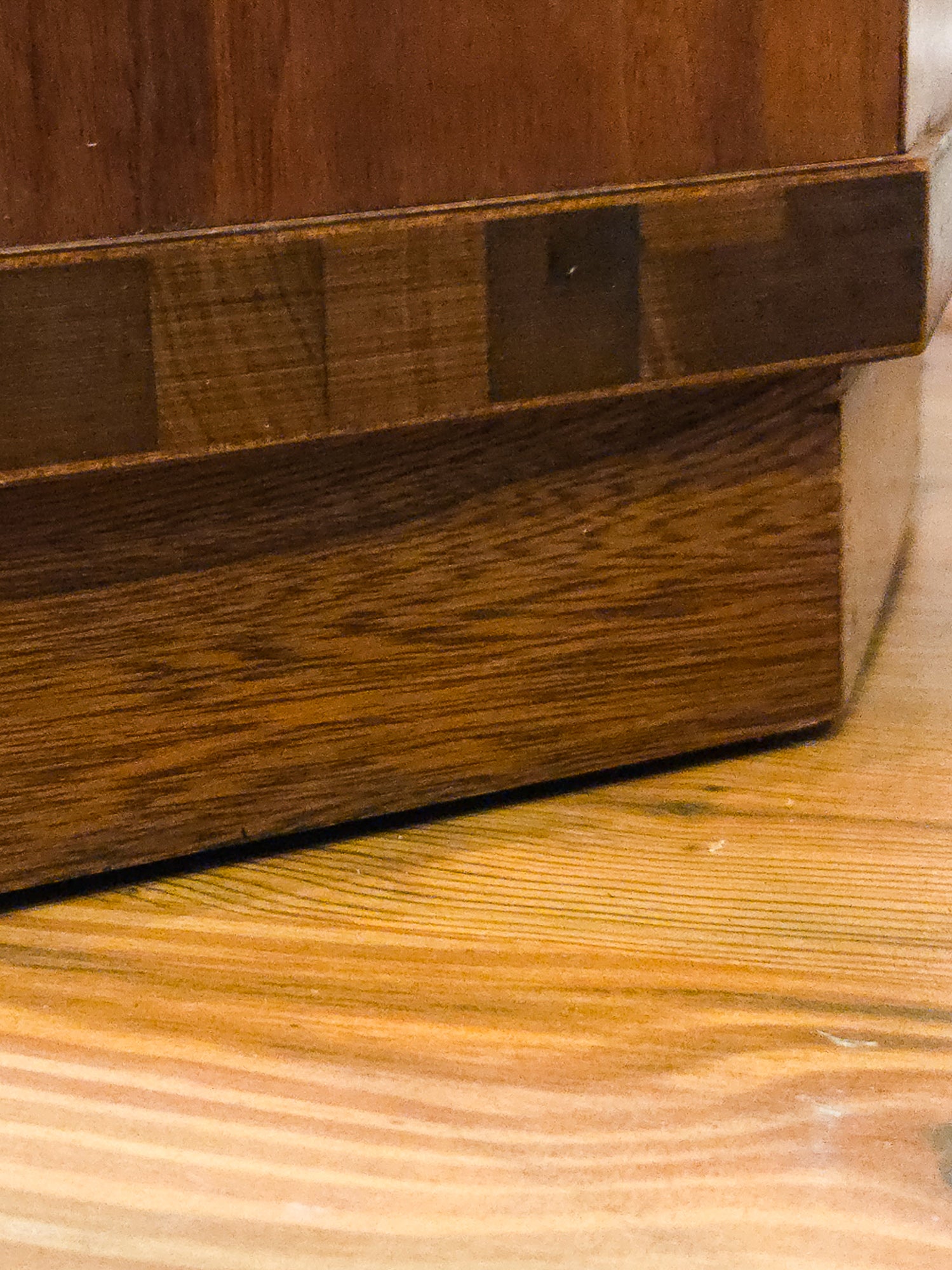 Close-up of a dark brown wooden furniture piece, showing a detailed wood-grain patterned apron and a solid wood base, set against a light wood floor.